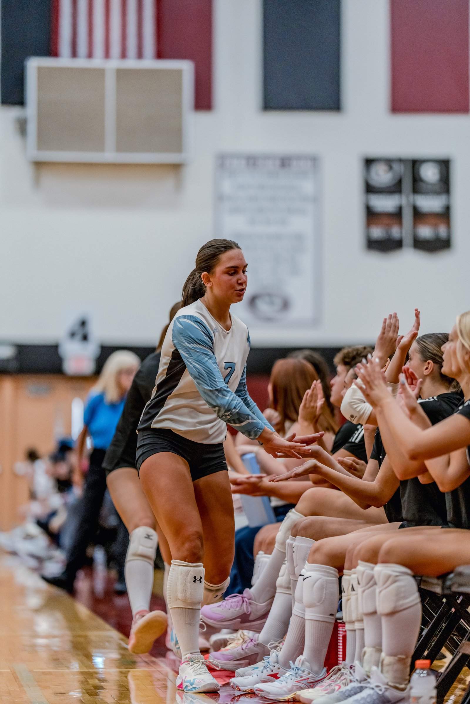 A player in a white and blue jersey with number 7 high-fives teammates in black jerseys on the bench during a volleyball game.