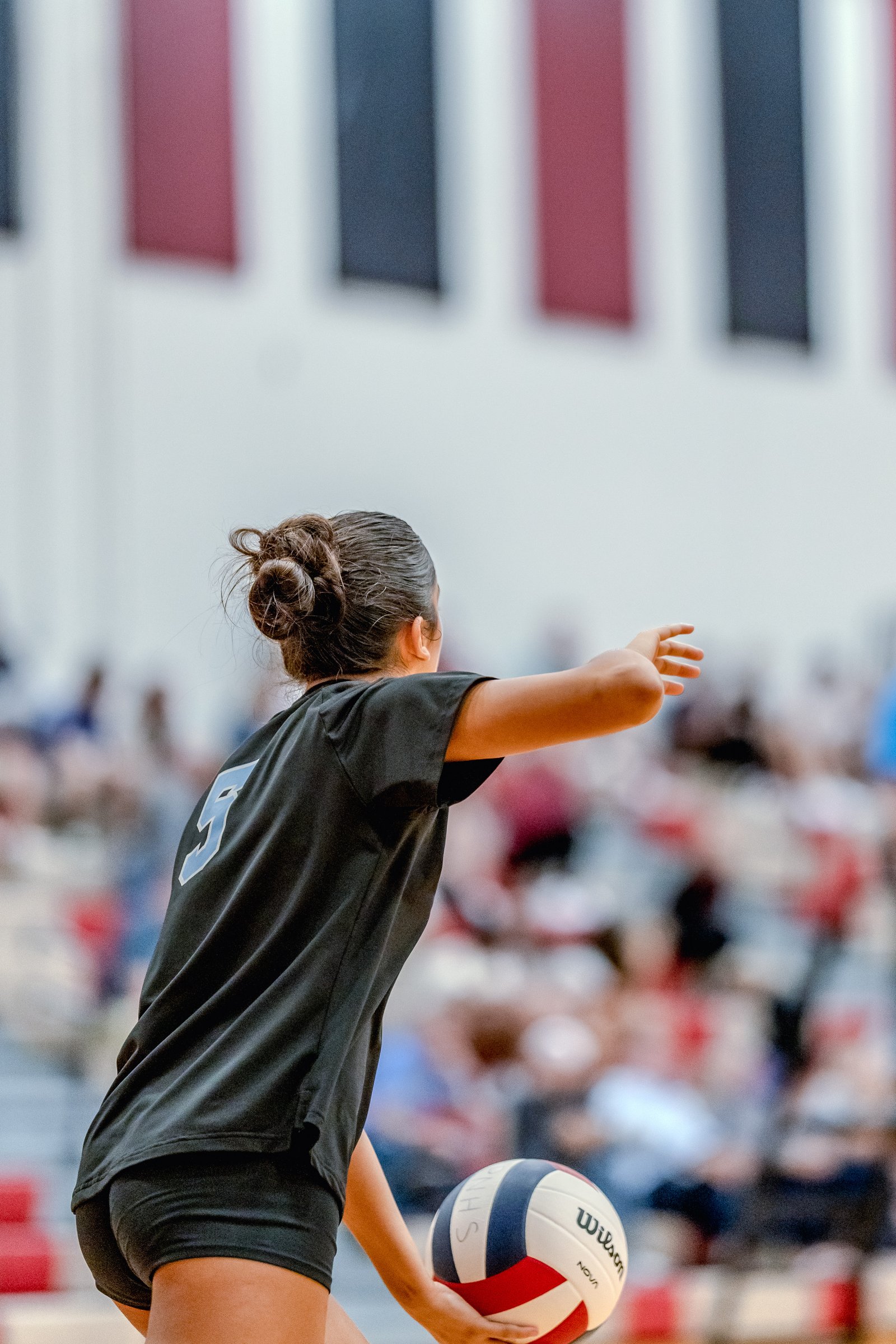 A player in a black jersey with a light blue number 5 prepares to serve a volleyball in a gymnasium with spectators.