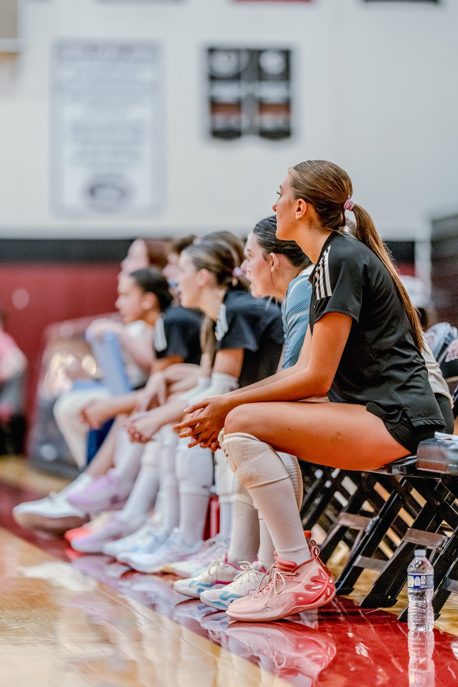 Players in black and blue jerseys sit on the bench, watching the game. A player in black has white stripes on her sleeve.