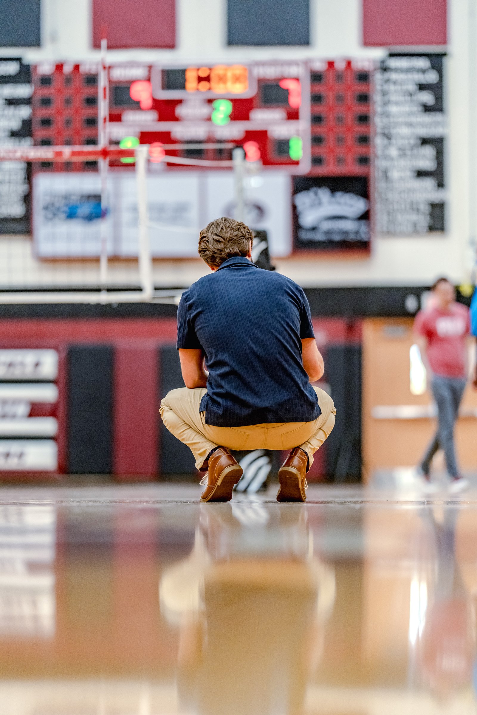 A player in a dark blue shirt and tan pants crouches on the court, facing away from the camera, with a blurred scoreboard in the background.