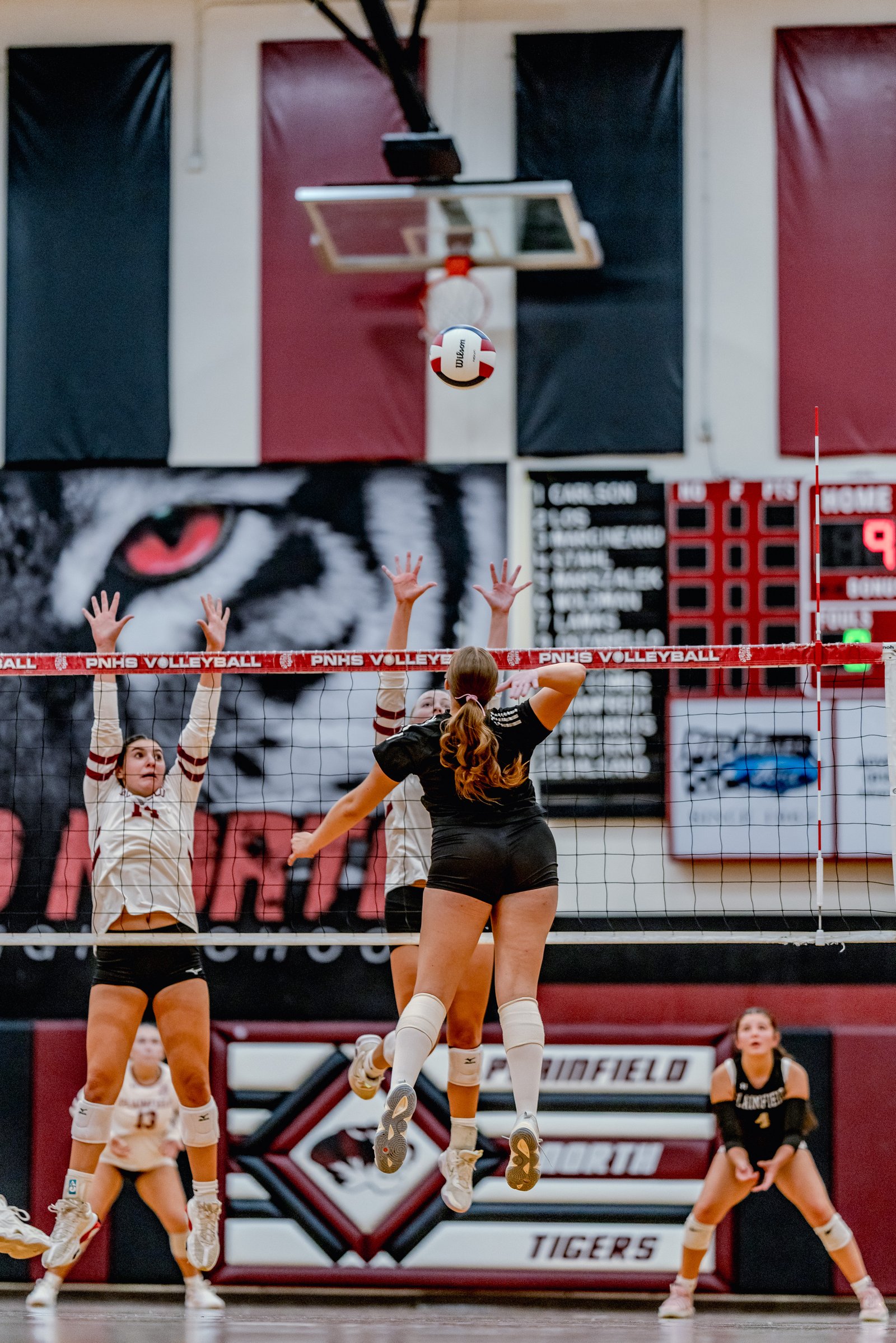 Players in white and black jerseys jump at the net during a volleyball game, with a player in white number 13 visible.
