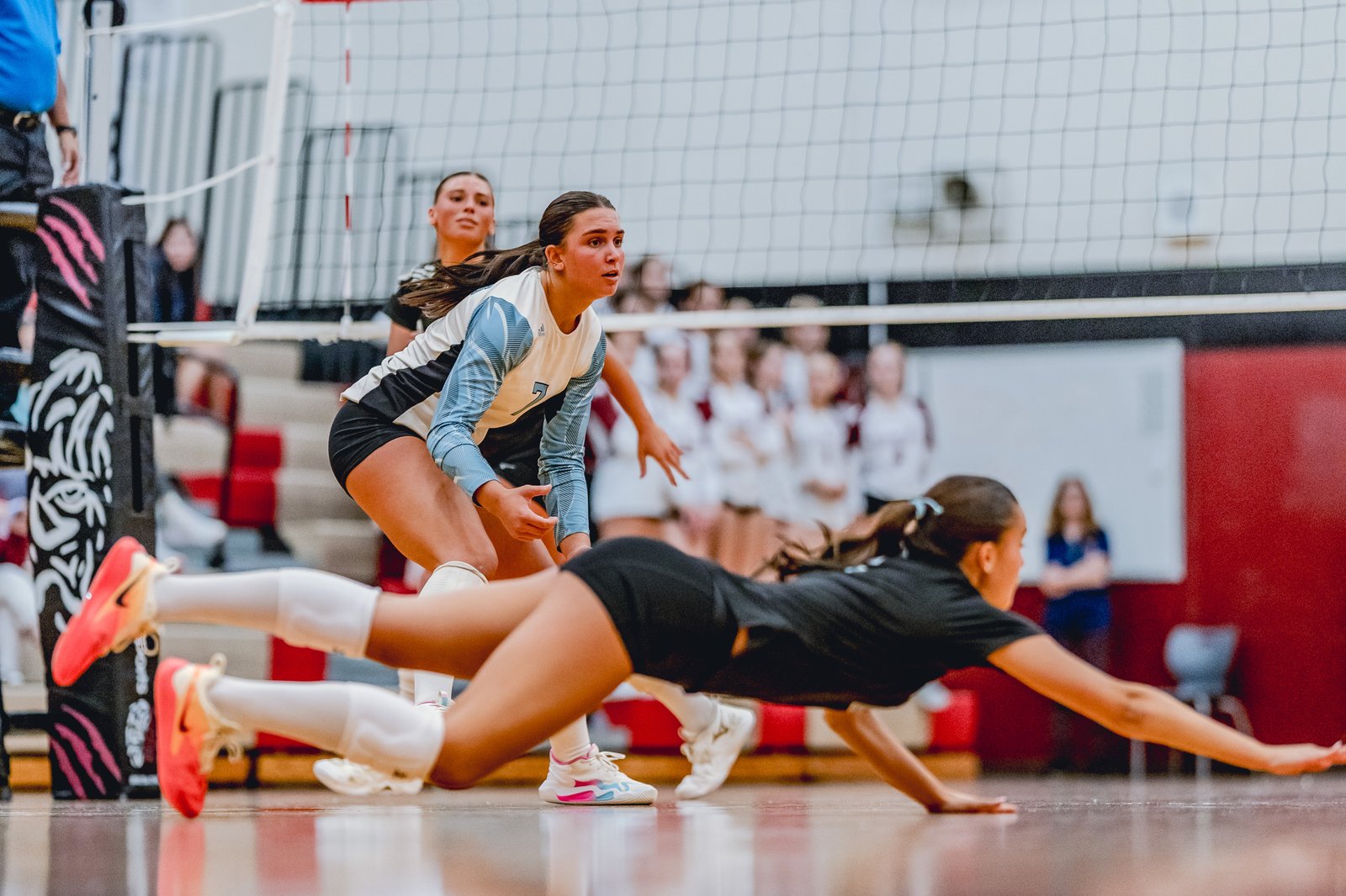 A player in black dives for a dig while a player in a light blue and white jersey prepares to play.