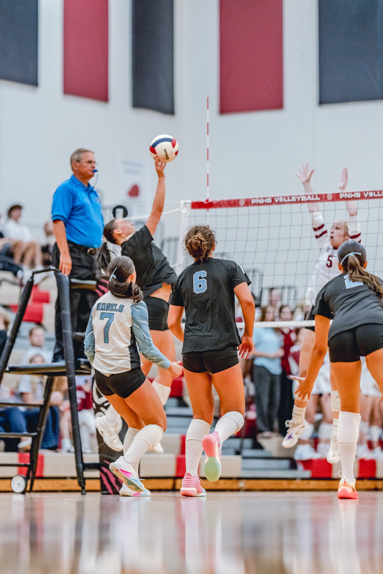A player in black jumps to hit the volleyball as players in light blue and white with maroon trim play defense during a game.