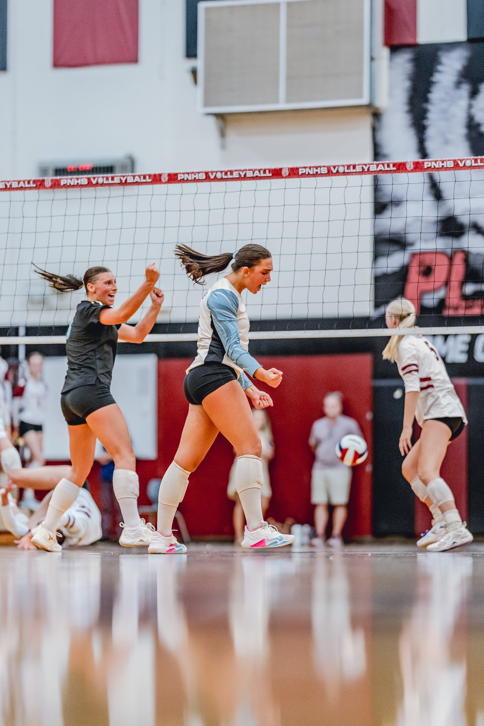 Players in black and light blue jerseys react during a volleyball game, with a player in a maroon jersey visible on the right.