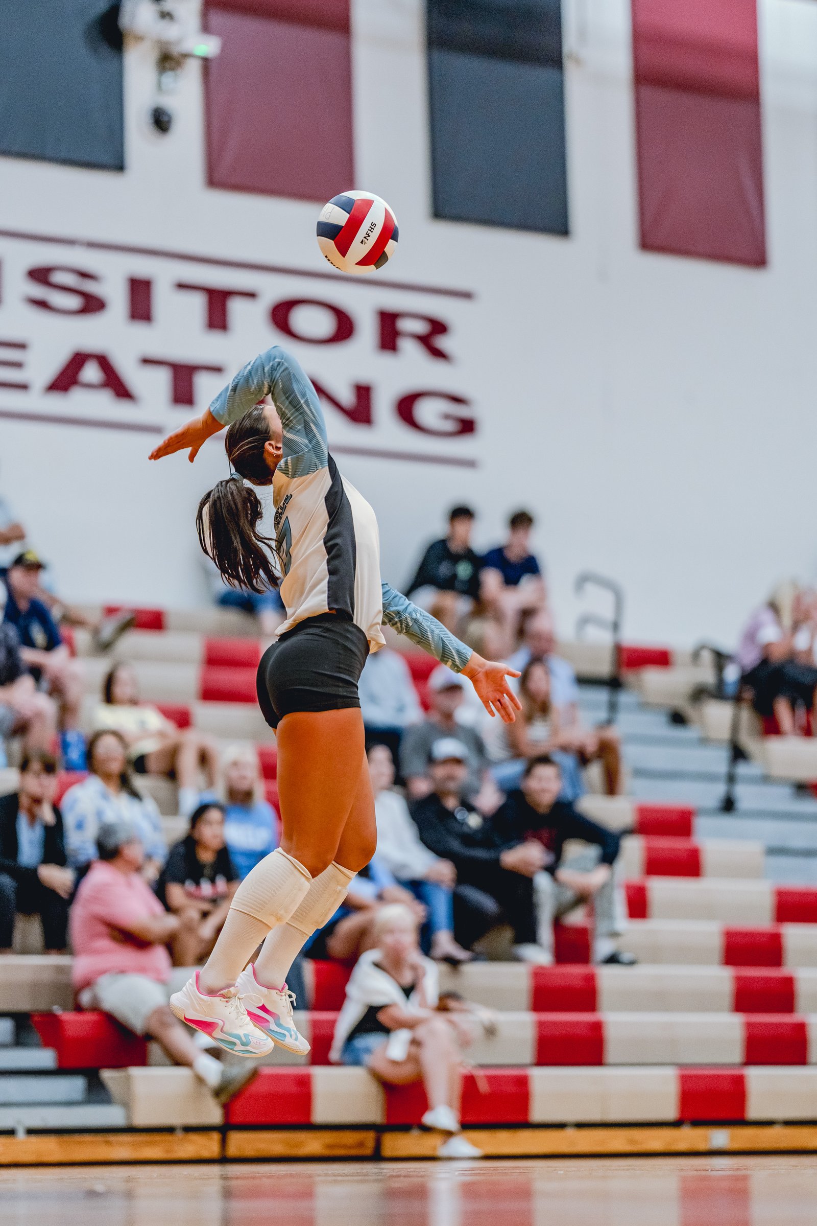 A player in a light blue and white jersey jumps to hit a volleyball during a game, with spectators in the background.