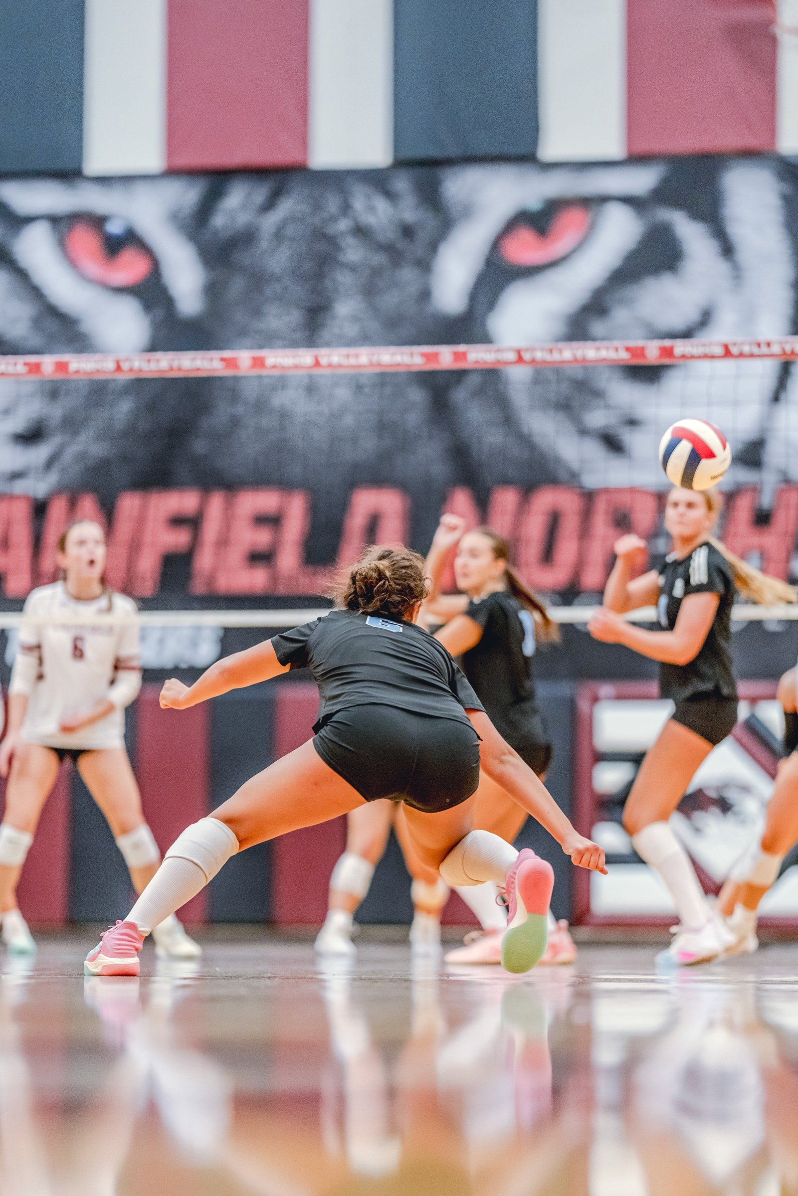 A player in black dives for a volleyball as teammates in black and white with maroon trim play on the court.