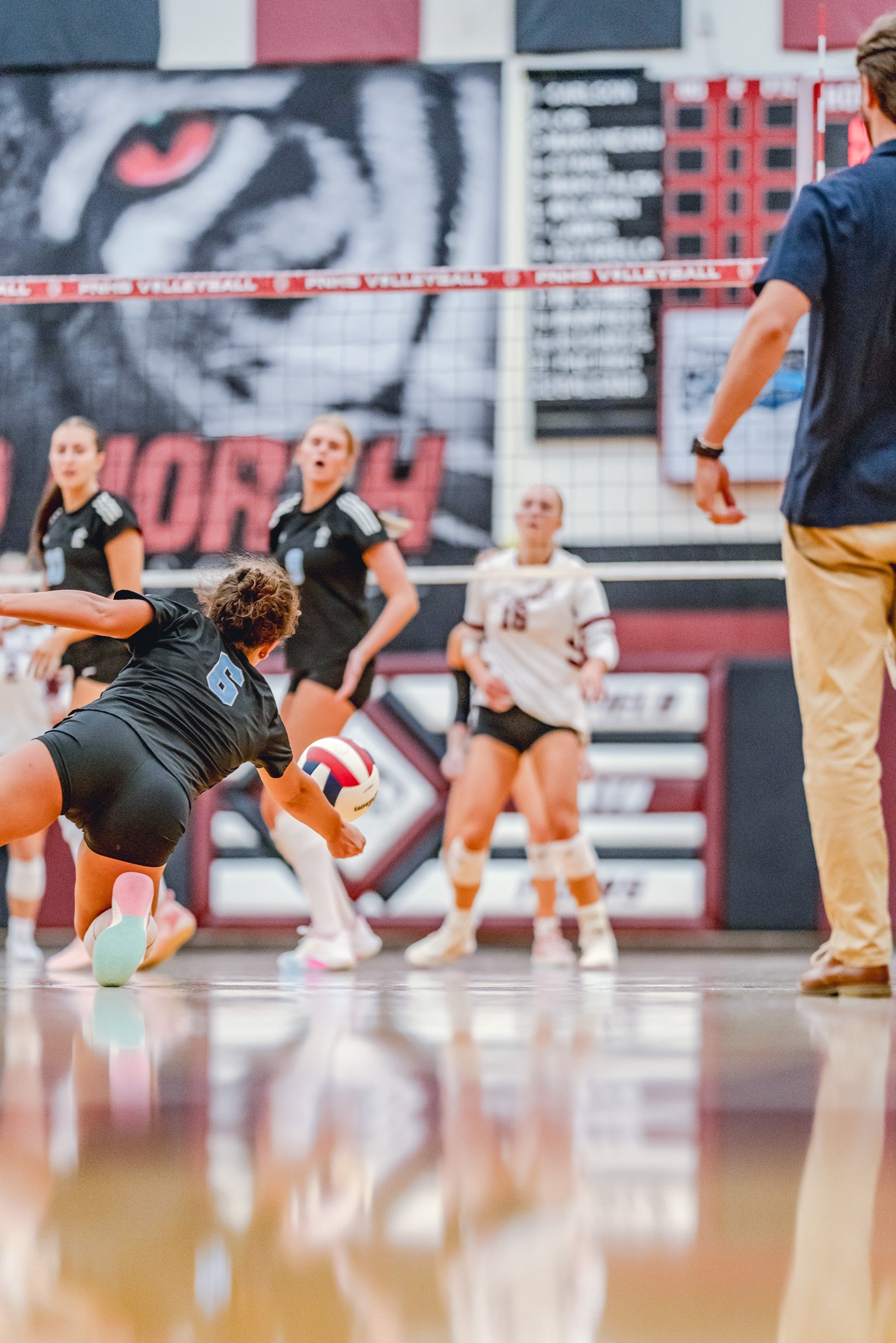 A player in a black top with blue number 6 dives for a volleyball, with teammates in black and white tops watching.