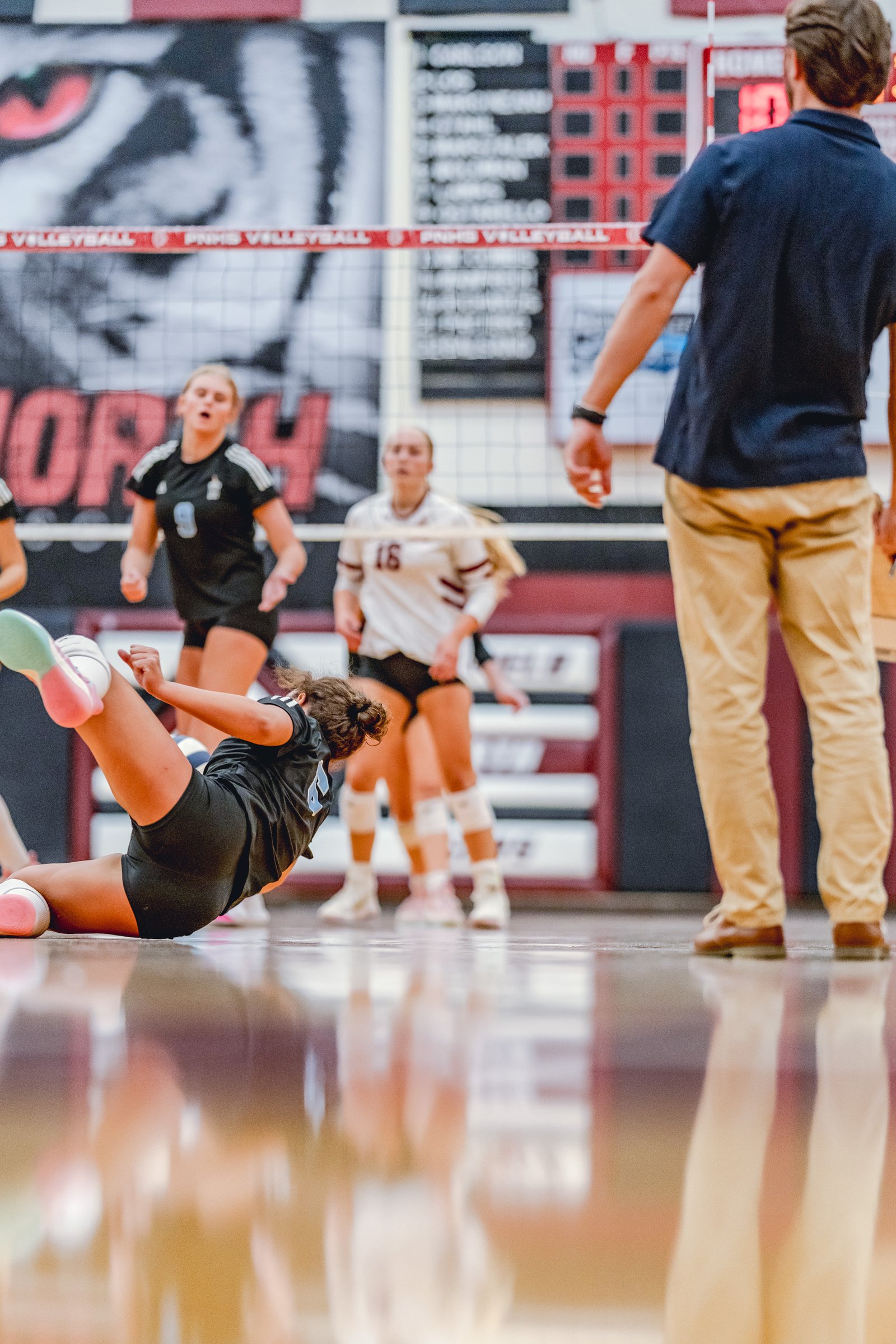 A player in black dives for a volleyball near the net, with teammates in black and white jerseys visible.