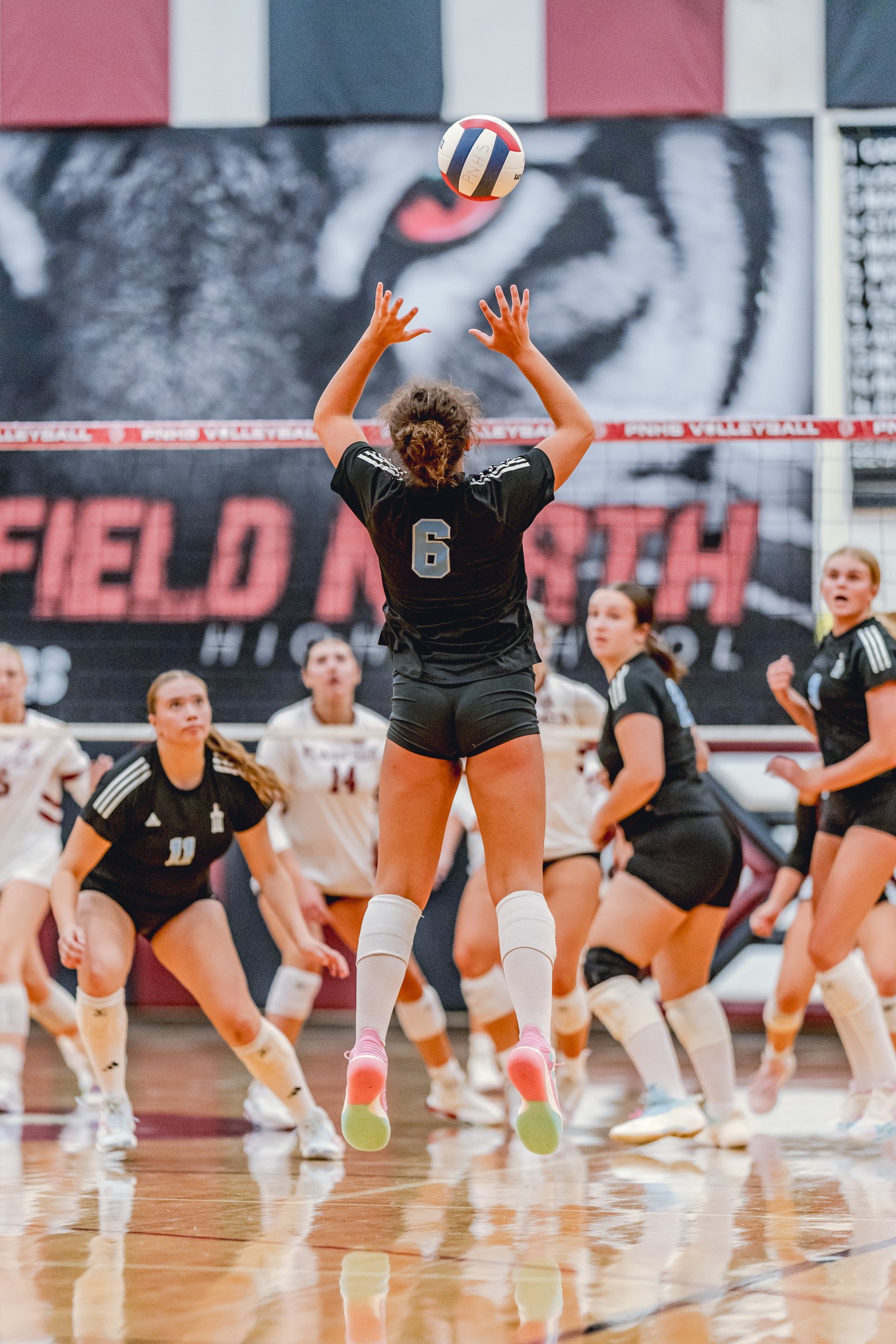 A player in black with jersey number 6 jumps to set the volleyball during a game against a team in white.