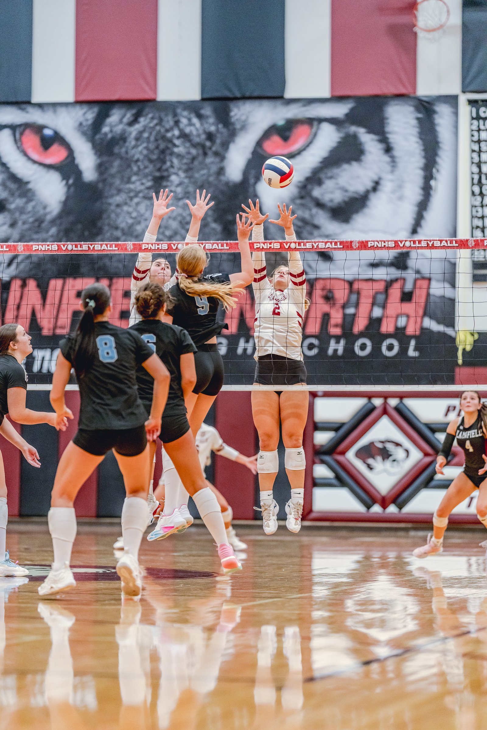 Players in black and white jerseys jump to block a volleyball over the net during a game.