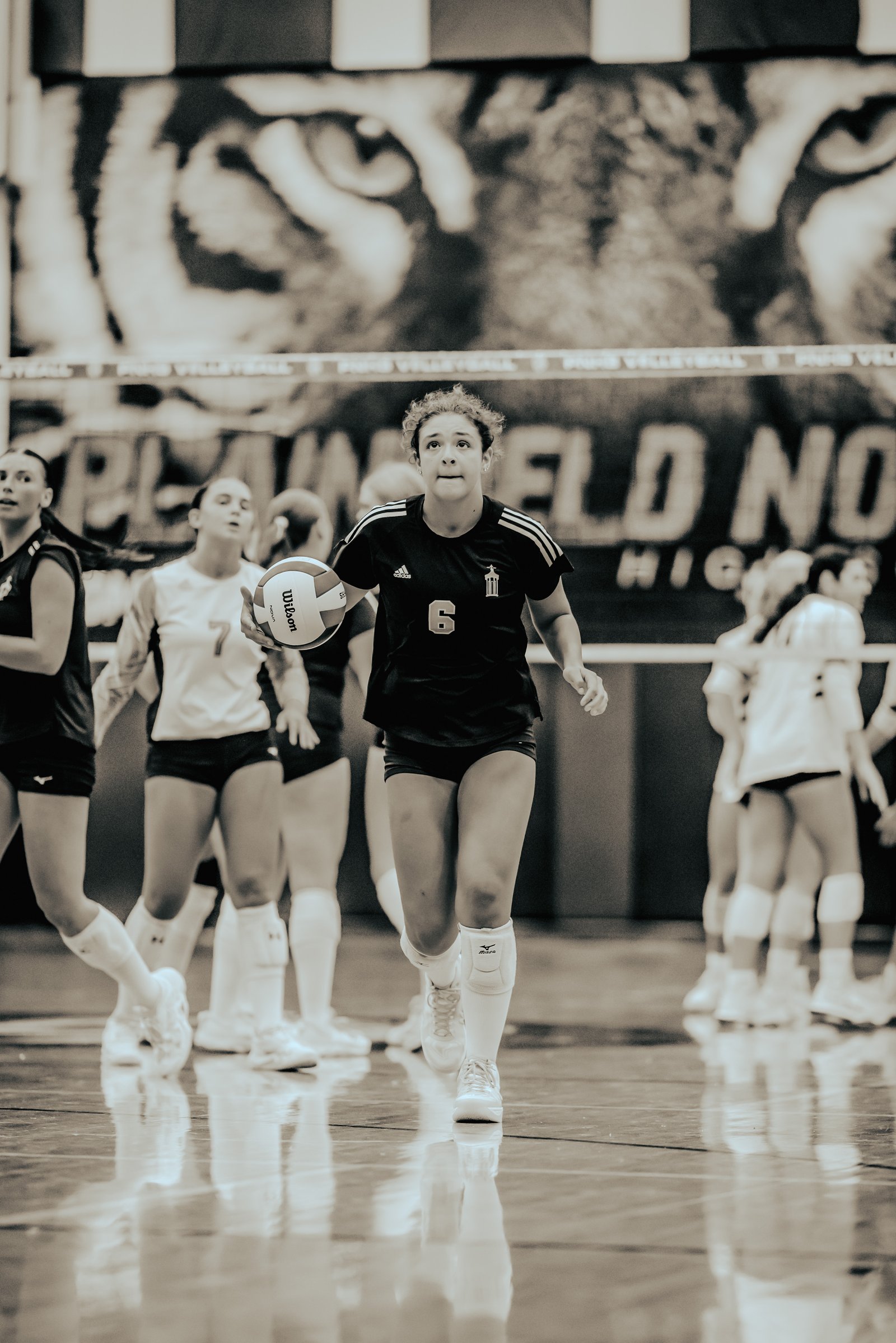 A player in a black top with number 6 holds a volleyball while other players in black and white uniforms are on the court.