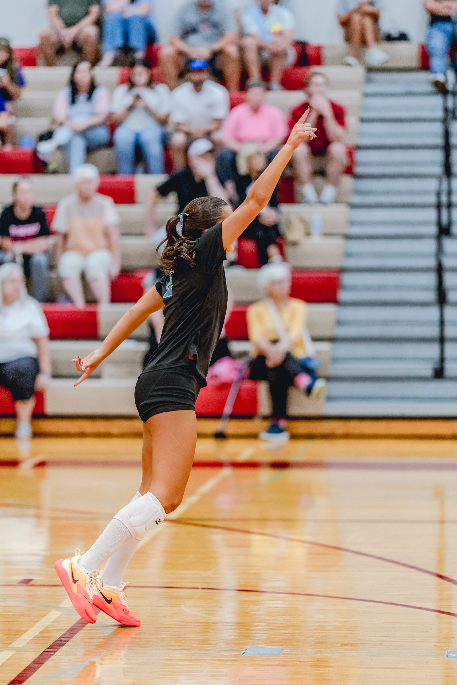 A player in a black top and shorts jumps to serve the volleyball in a gymnasium with spectators in the background.