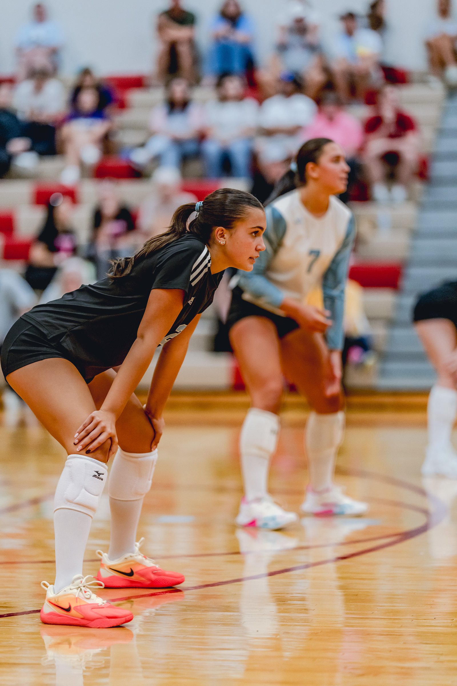A player in black crouches on the court, while a player in white and blue with jersey number 7 stands behind her during a volleyball game.