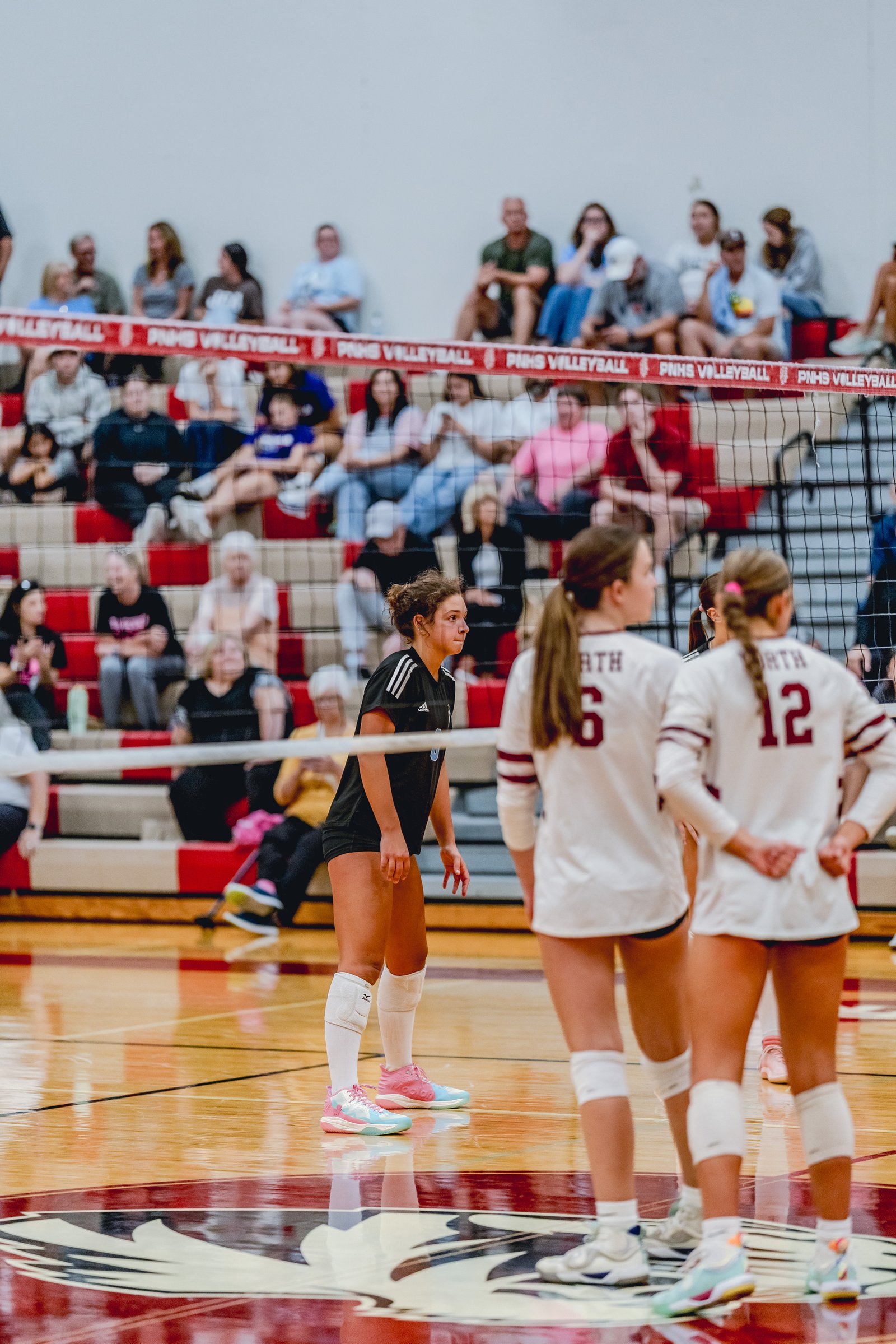 A player in a black top and shorts stands on the court, while players in white jerseys with maroon trim are in the foreground during a volleyball game.