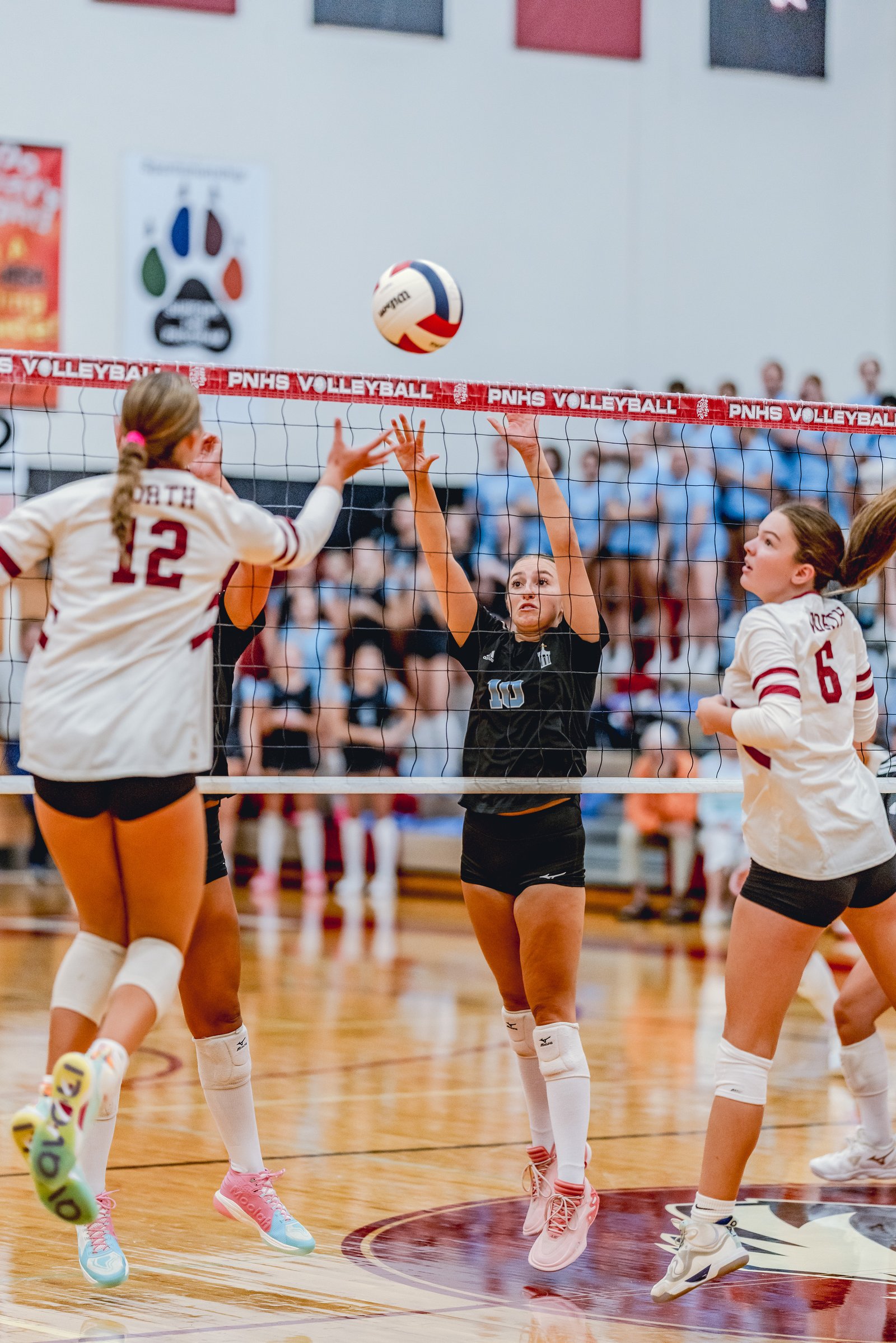 Players in white and black jerseys jump at the net during a volleyball game, with a ball in the air.