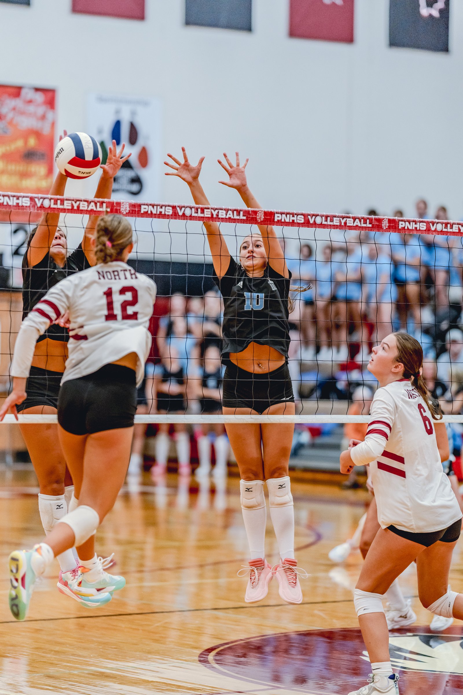 Players in white and black jerseys jump at the net during a volleyball game, with one player in white wearing number 12 and another in black wearing number 10.