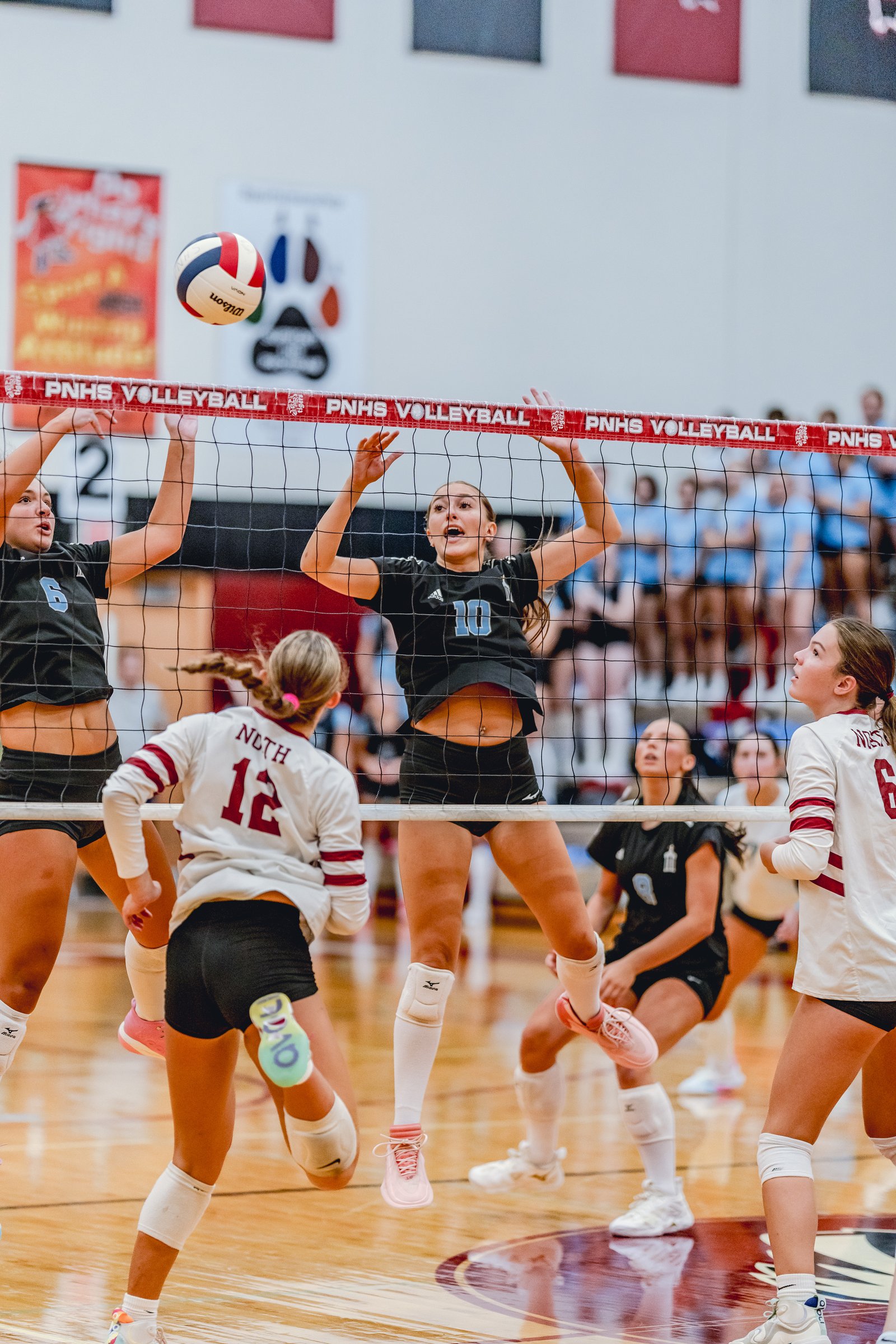 Players in black and white jerseys jump at the net during a volleyball game, with a player in black jersey number 10 reaching for the ball.