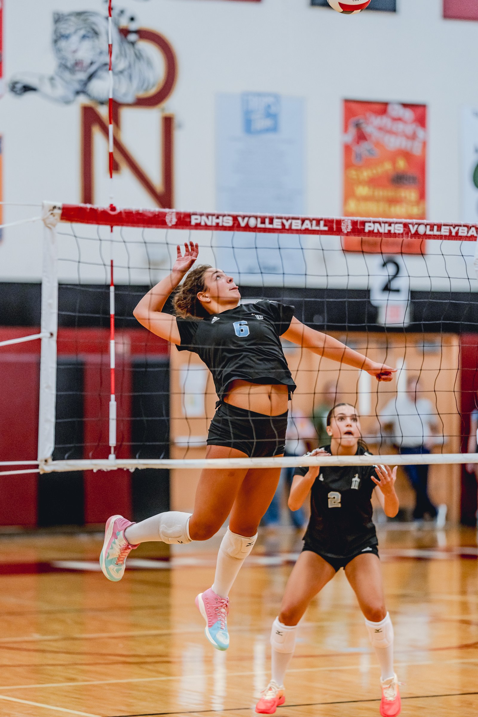 A player in a black top with number 6 jumps to hit a volleyball, with another player in a black top watching.
