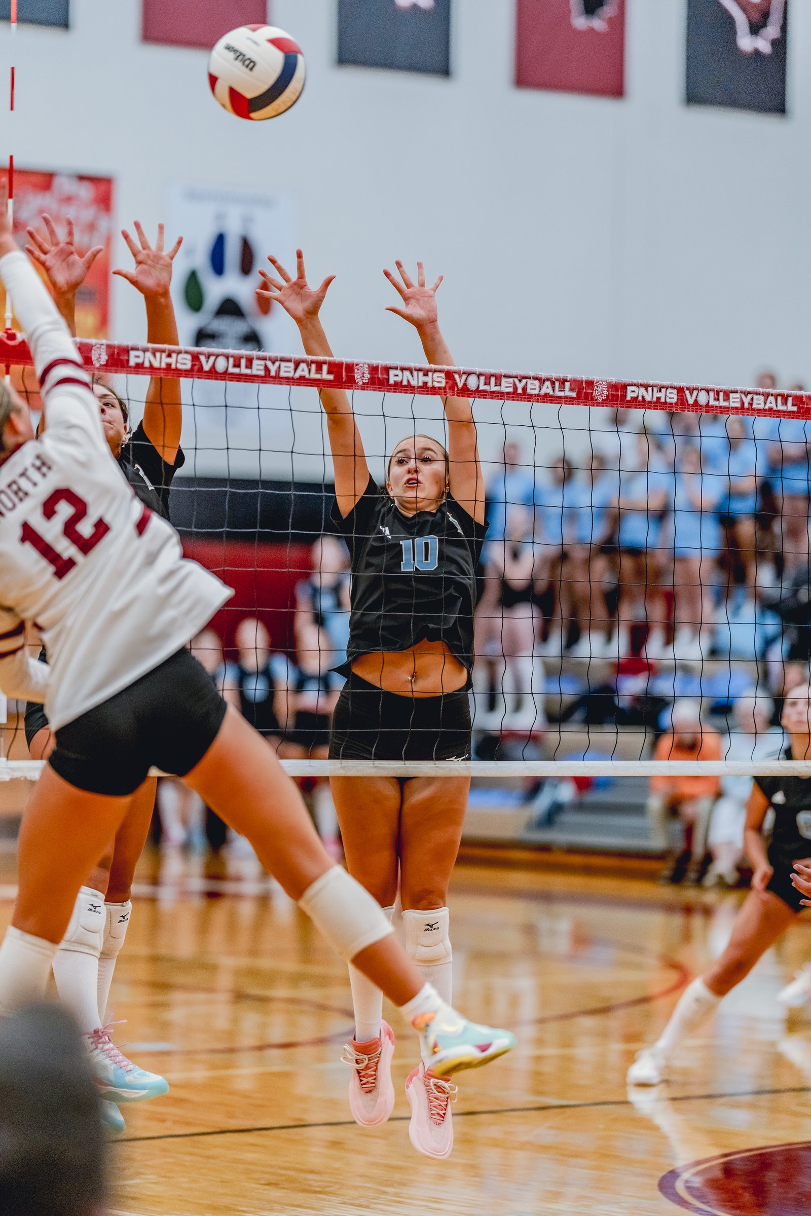 Players in black and white jerseys jump at the net during a volleyball game, with a ball in the air.