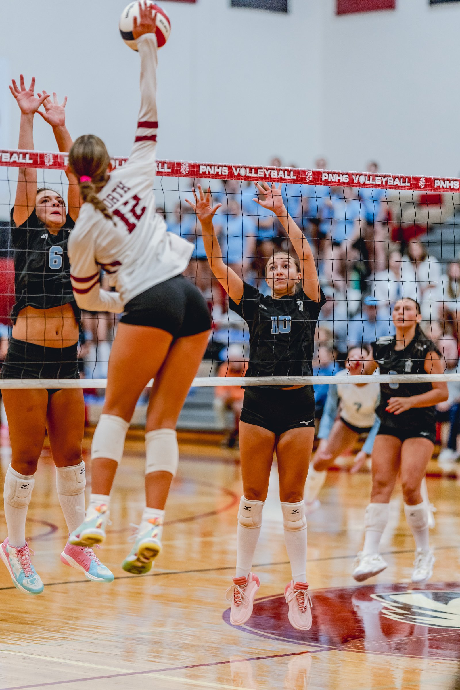 A player in white with maroon stripes spikes the volleyball over the net as players in black with blue numbers attempt to block.