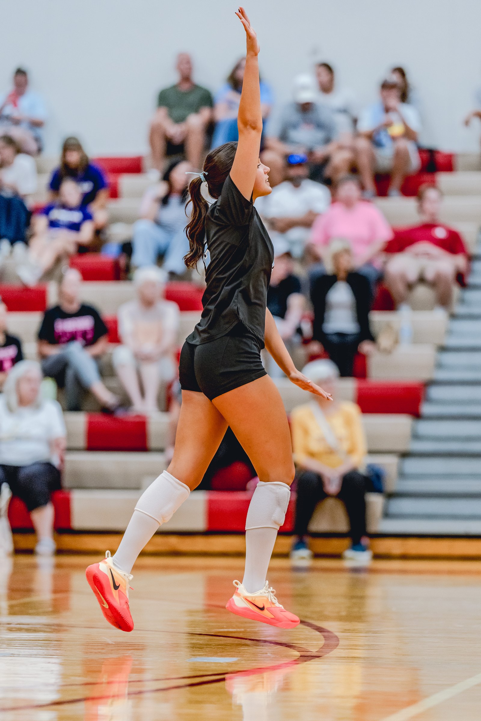 A player in a black top and shorts jumps with her arm raised, white socks, and pink shoes on a wooden court with spectators in the background.