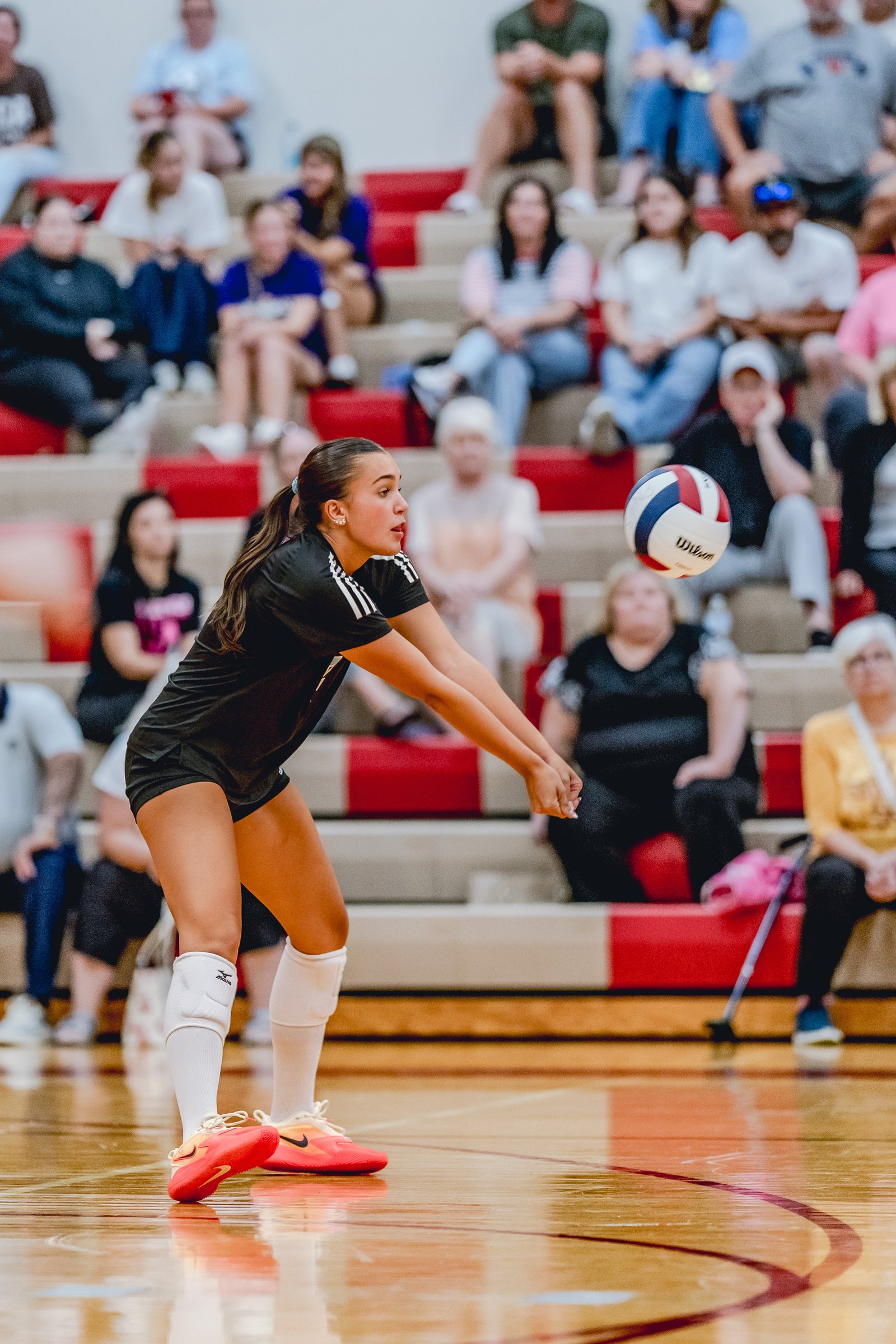 A player in a black top and shorts passes a volleyball on the court with spectators in the background.