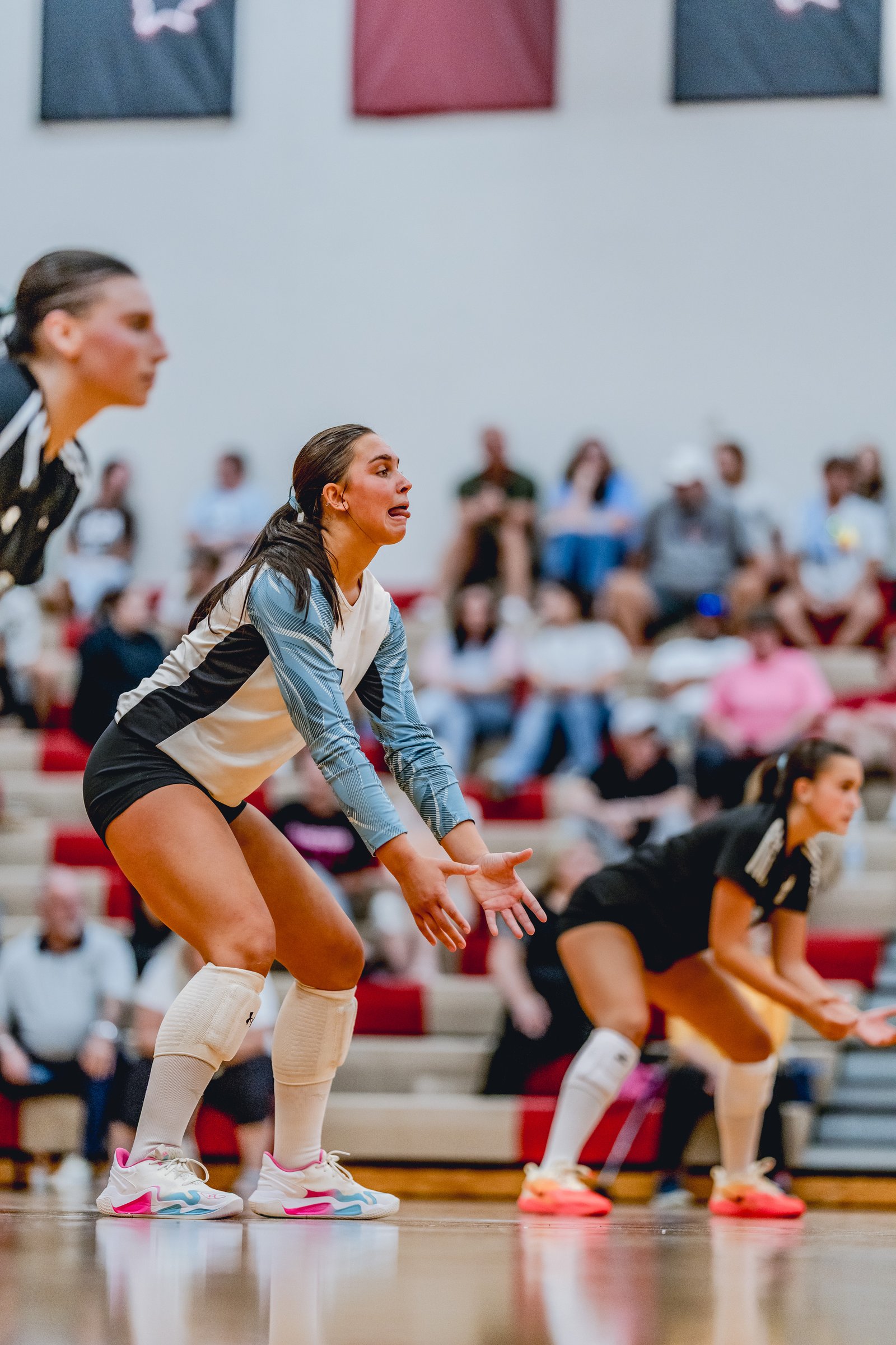 A player in a light blue and white jersey prepares to receive a volleyball on the court with spectators in the background.
