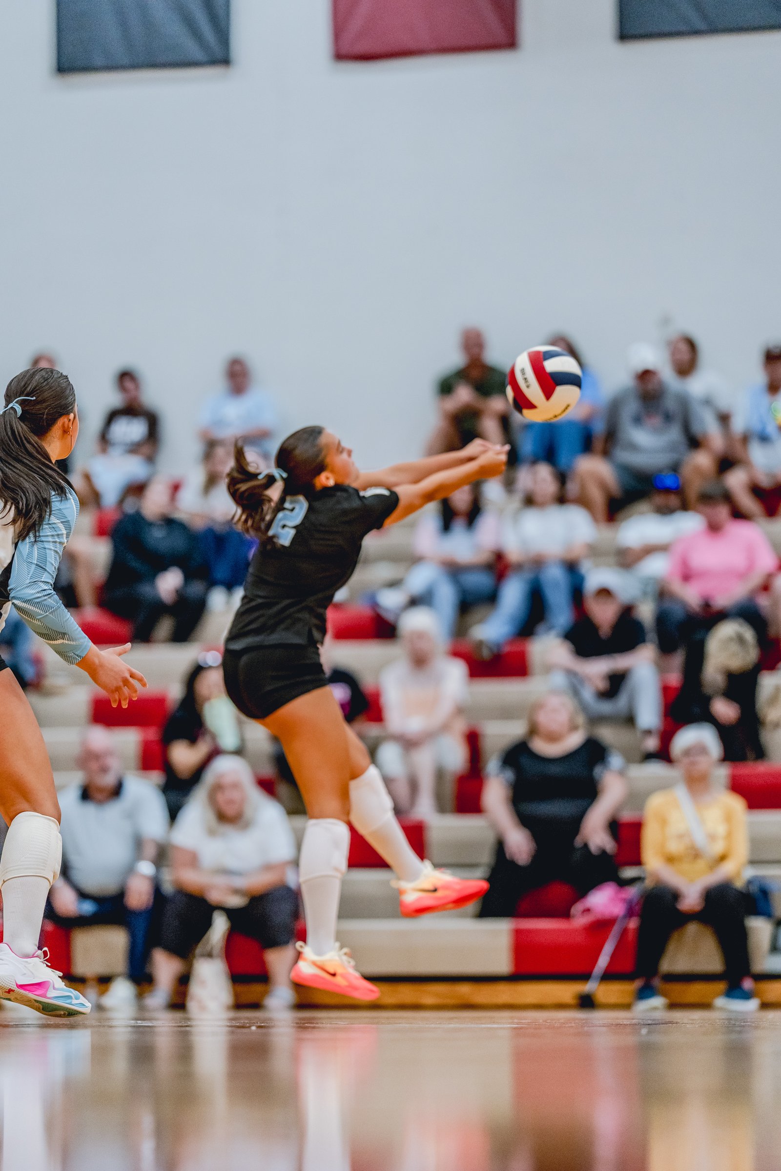 A player in a black top with jersey number 2 passes a volleyball during a game, with spectators in the background.