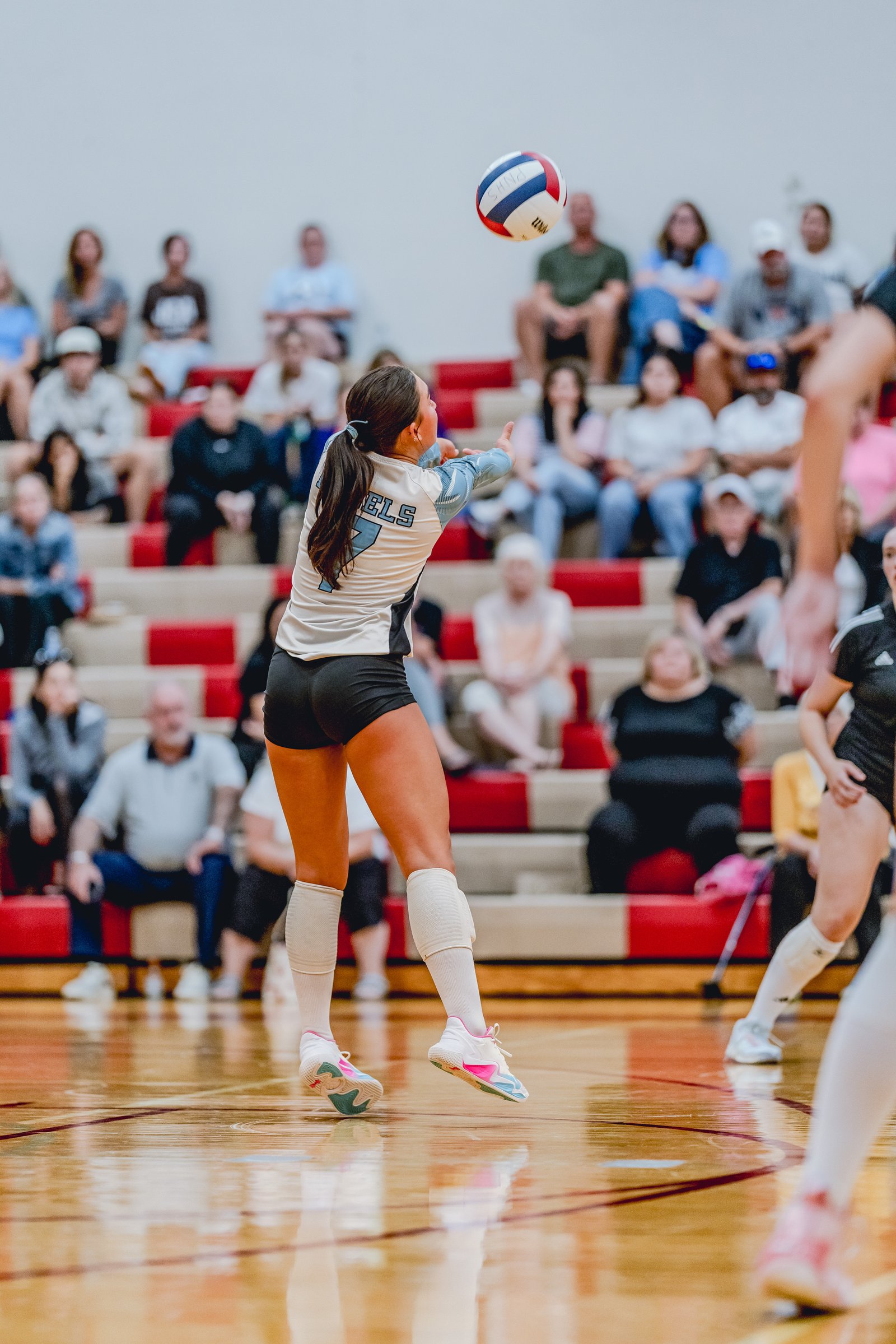 A player in a light blue and white jersey with number 7 sets the volleyball during a game with spectators in the background.