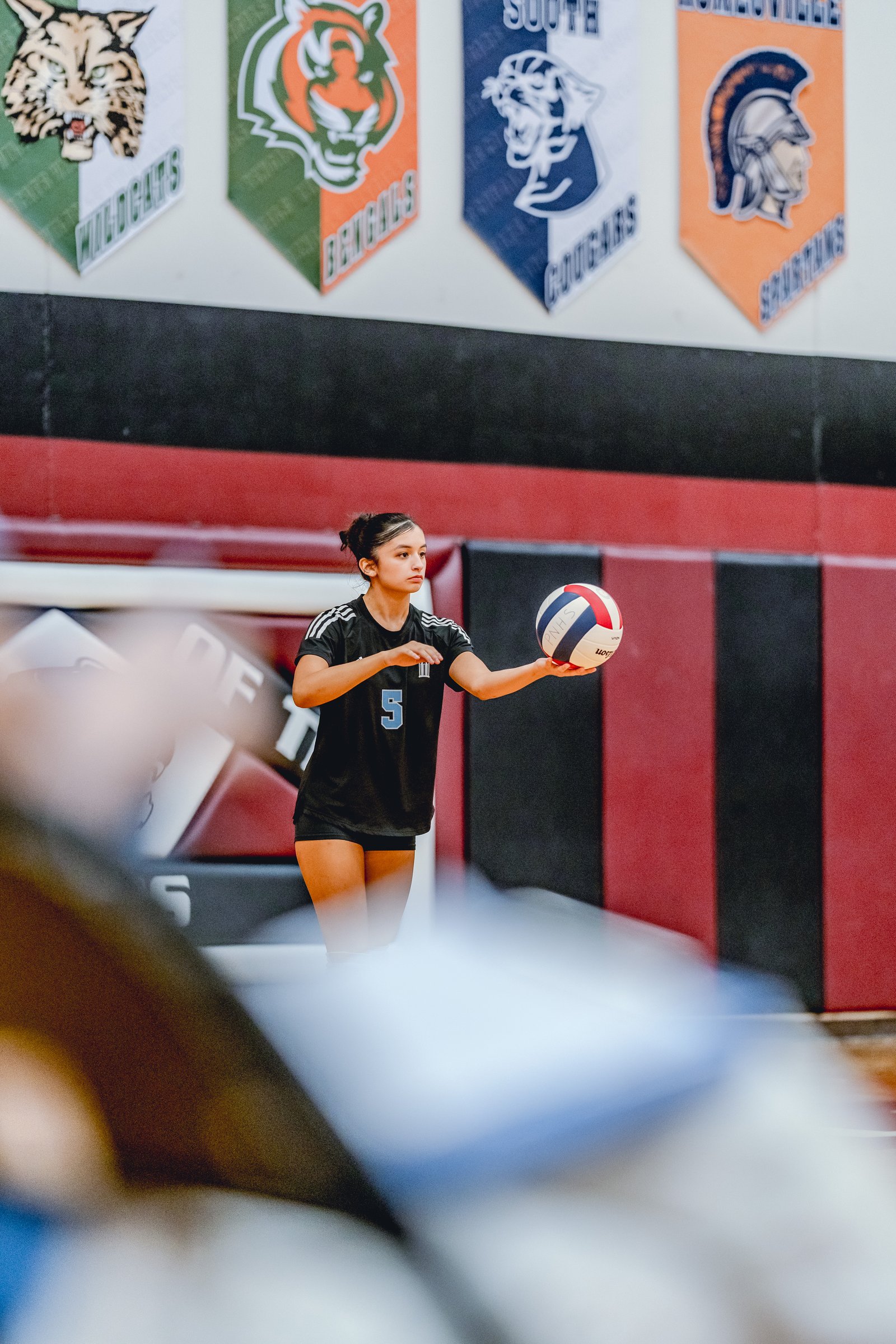 A player in a black jersey with white stripes and number 5 prepares to serve a volleyball indoors.