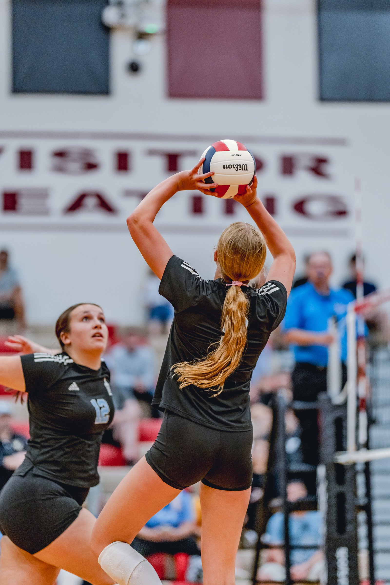 A player in black prepares to set a volleyball, with another player in black and number 12 visible on the court.