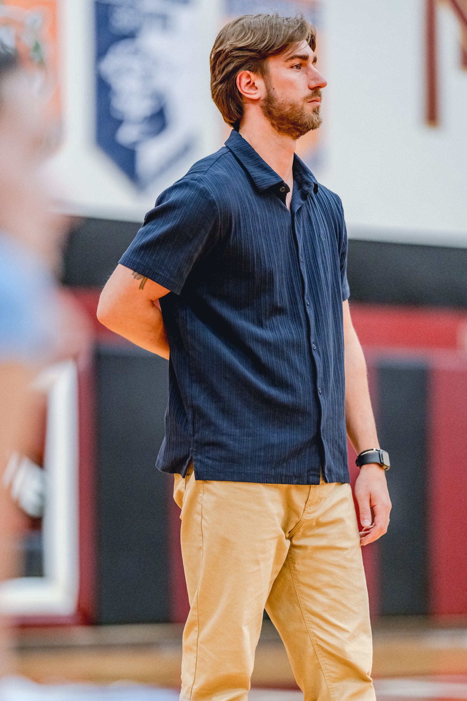 A player in a navy shirt and tan pants stands on a court with blurred players in the background.