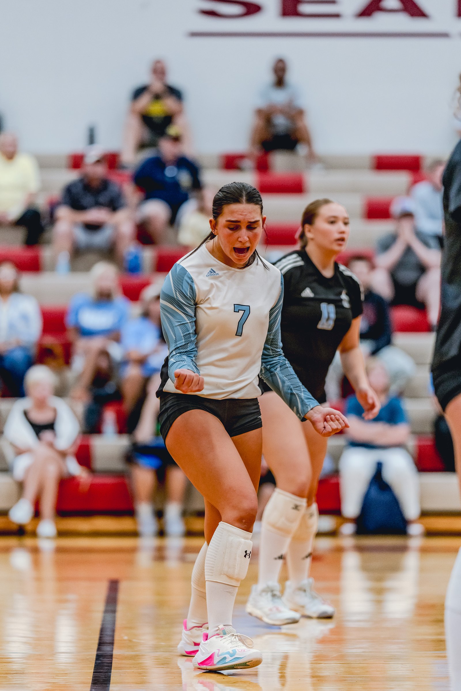 A player in a white and blue jersey with number 7 yells during a volleyball game, with a player in a black jersey behind her.