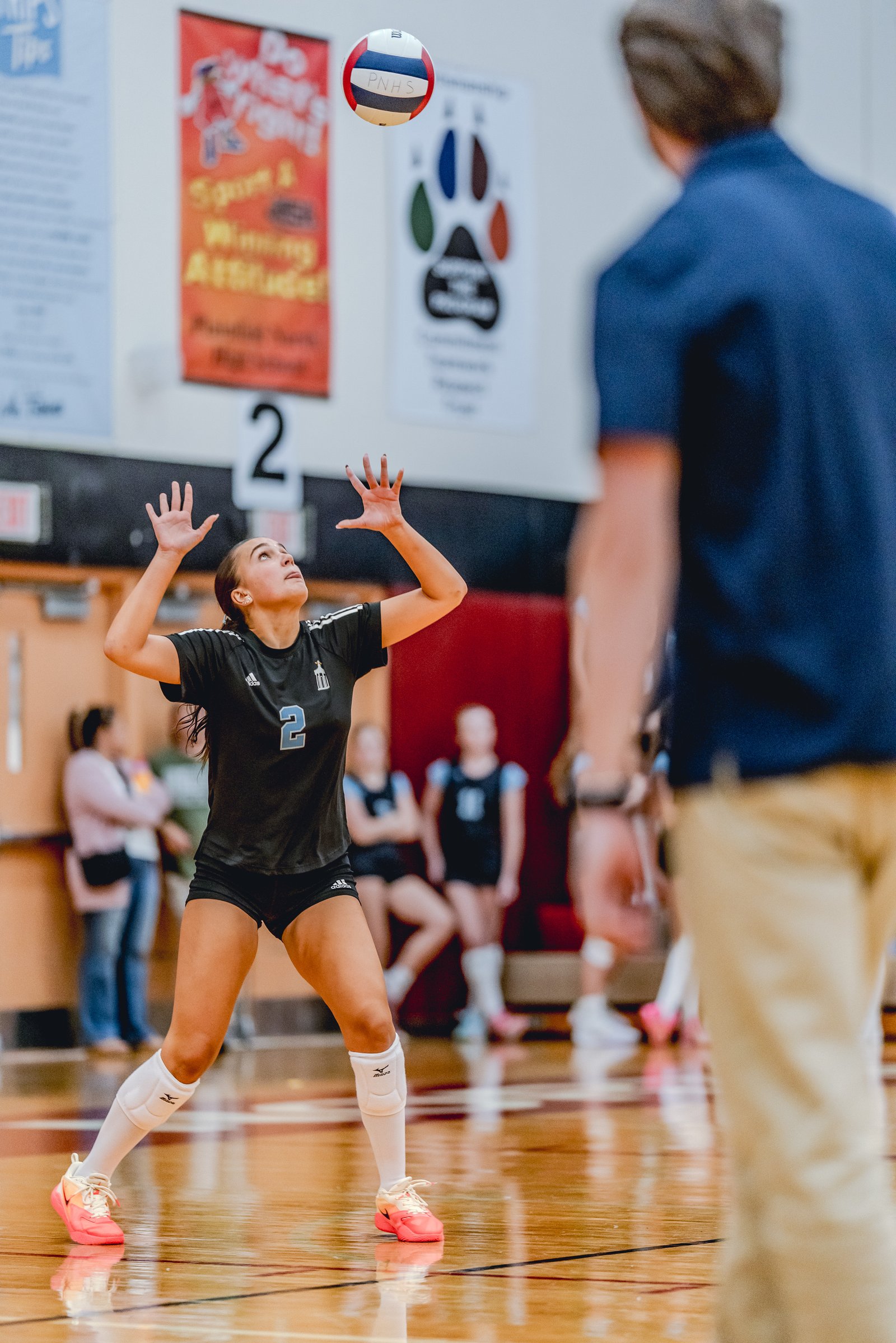 A player in a black top with number 2 prepares to receive a volleyball in a gymnasium.