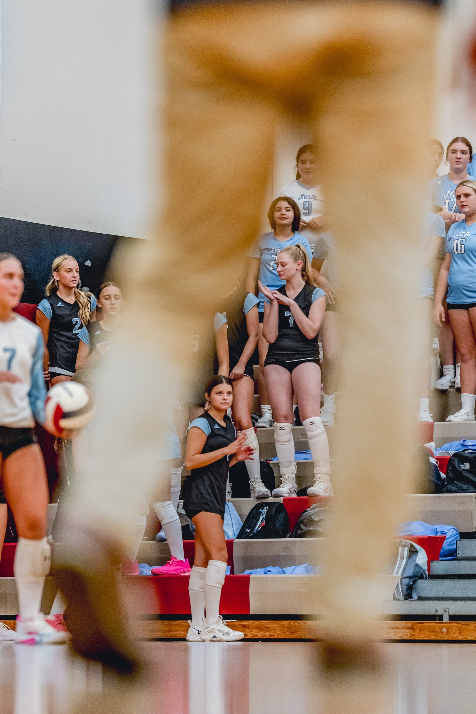 Players in light blue and black jerseys on bleachers, one player in black with jersey number 7 holds a volleyball.
