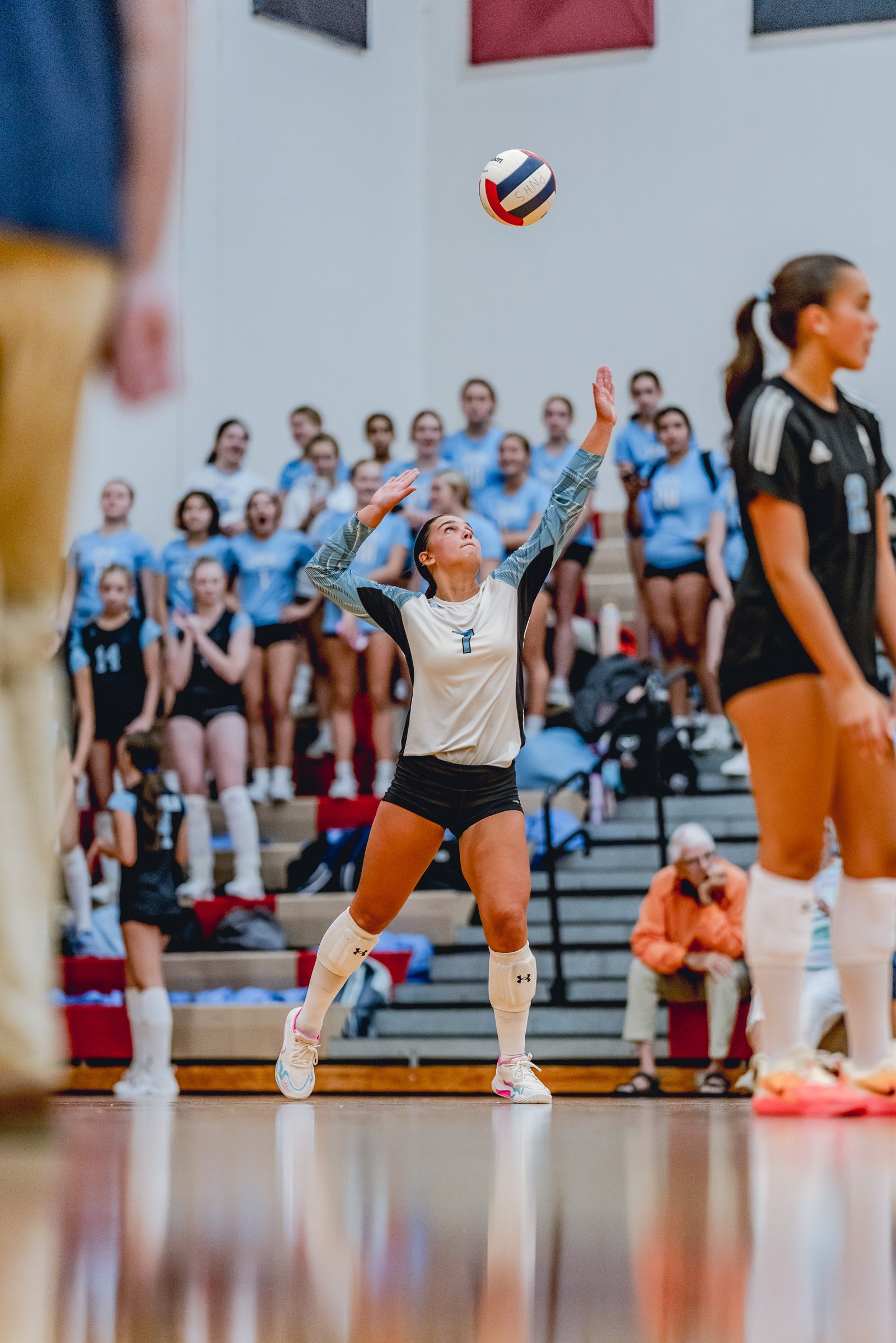 A player in a white and blue jersey serves a volleyball during a game, with teammates in blue and black jerseys watching from the bleachers.