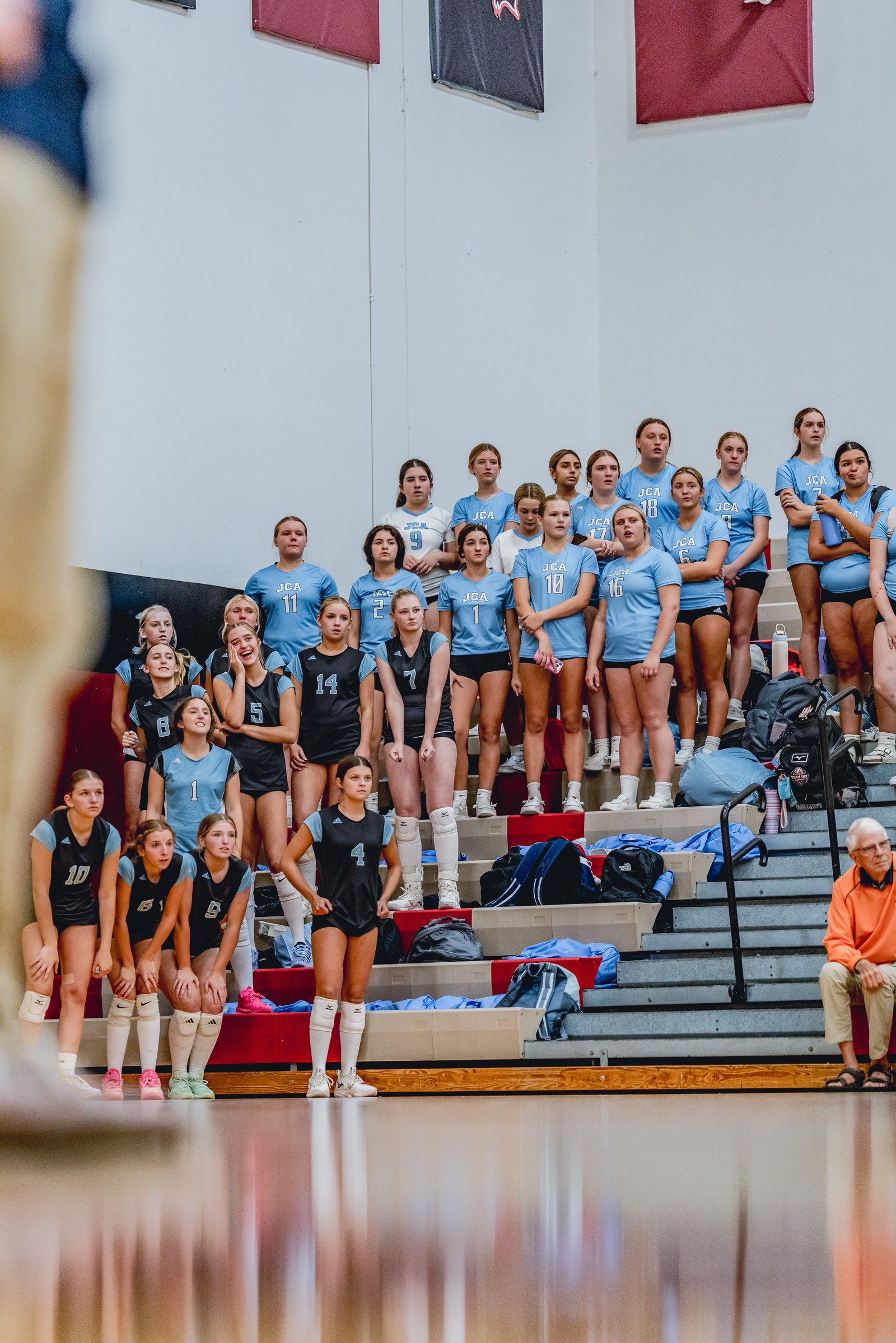 Players in light blue and black jerseys stand on bleachers. Visible jersey numbers include 10, 5, 14, 7, 4, 1, 11, 9, 2, 18, 17, 6, 16.