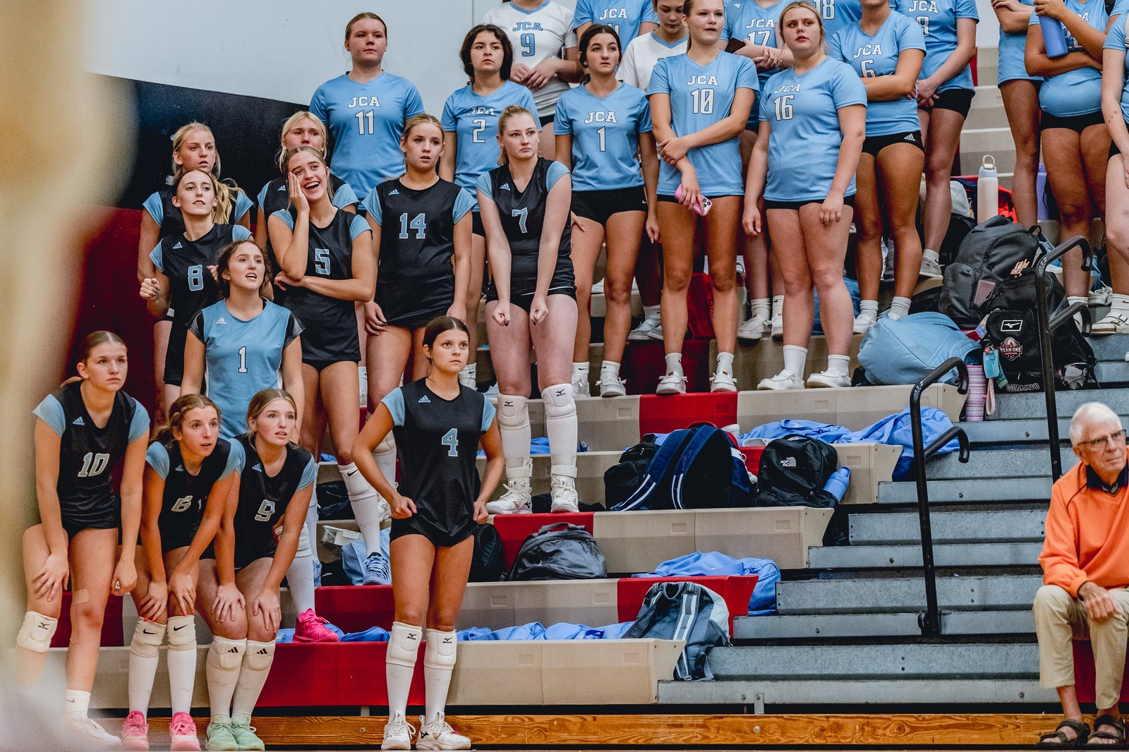 Players in light blue and black jerseys watch the game from the bleachers, with one player in a black jersey number 4 in the foreground.