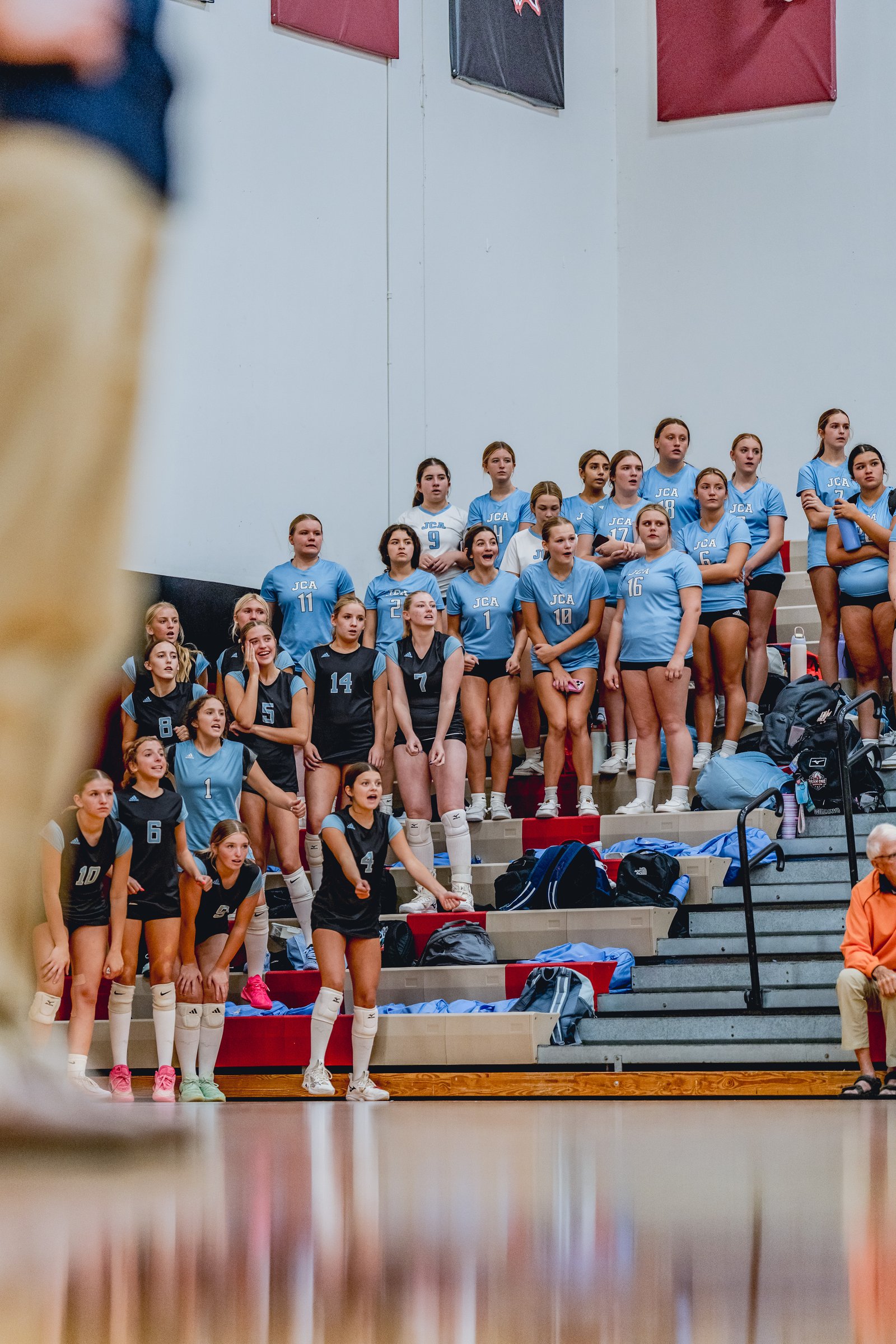 Players in light blue and black jerseys watch the game from the bleachers, with one player in black and blue in the foreground gesturing.