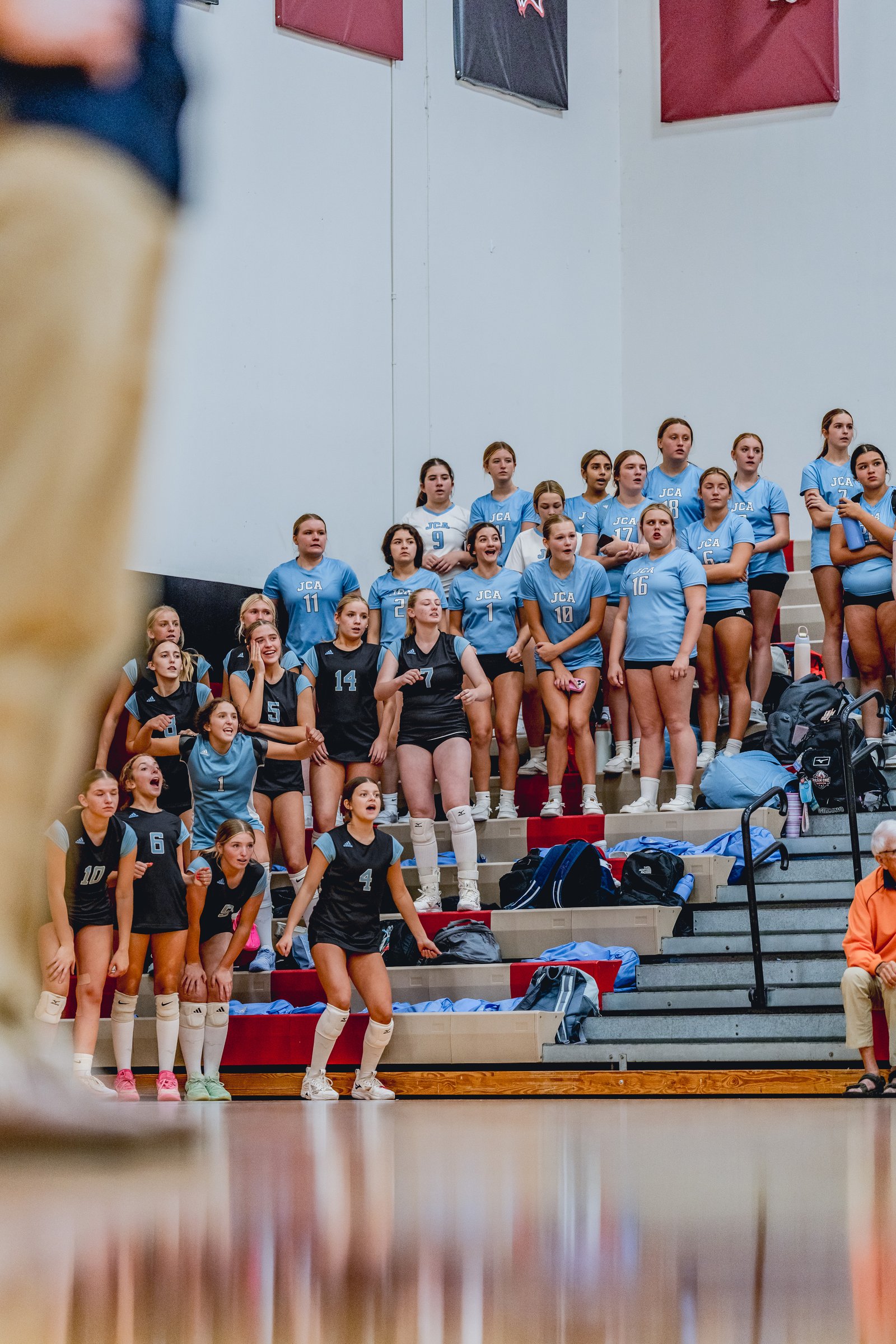 Players in light blue and black jerseys watch the game from the bleachers, with one player in black in the foreground.