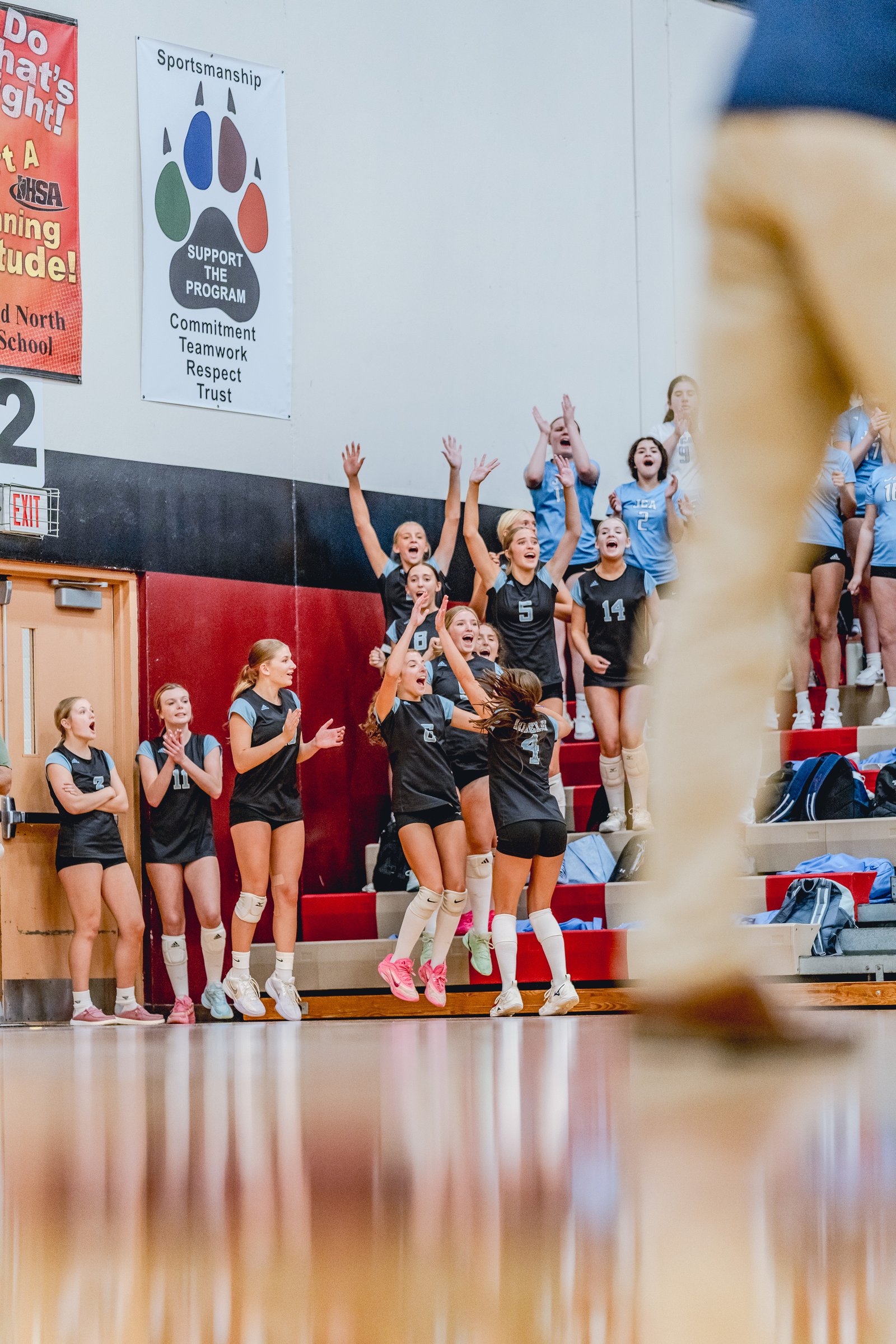 Players in black and light blue jerseys cheer on the court, with some jumping and raising their hands.