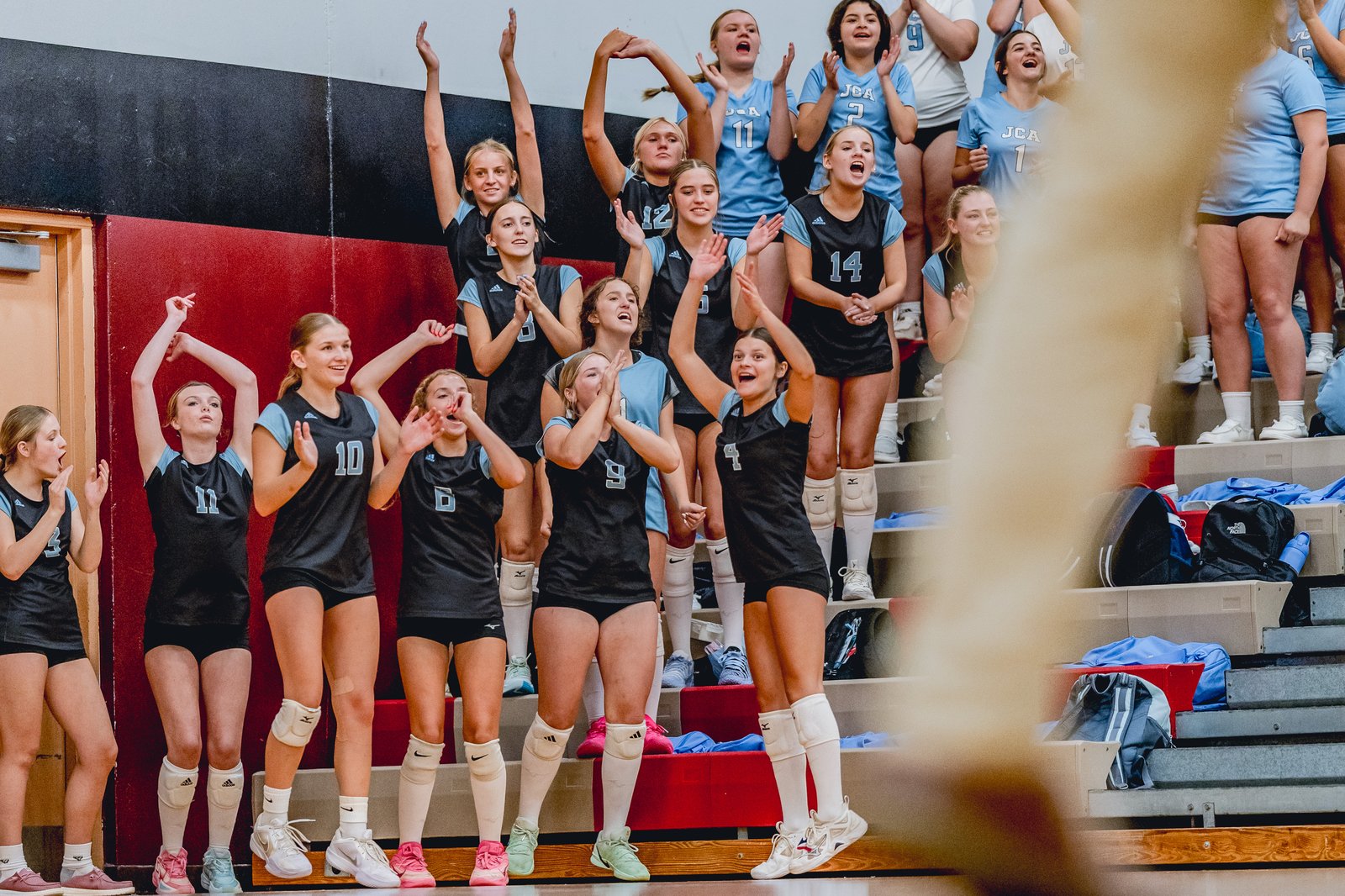 Players in black and light blue jerseys cheer from the bleachers, some with arms raised and hands clapping.