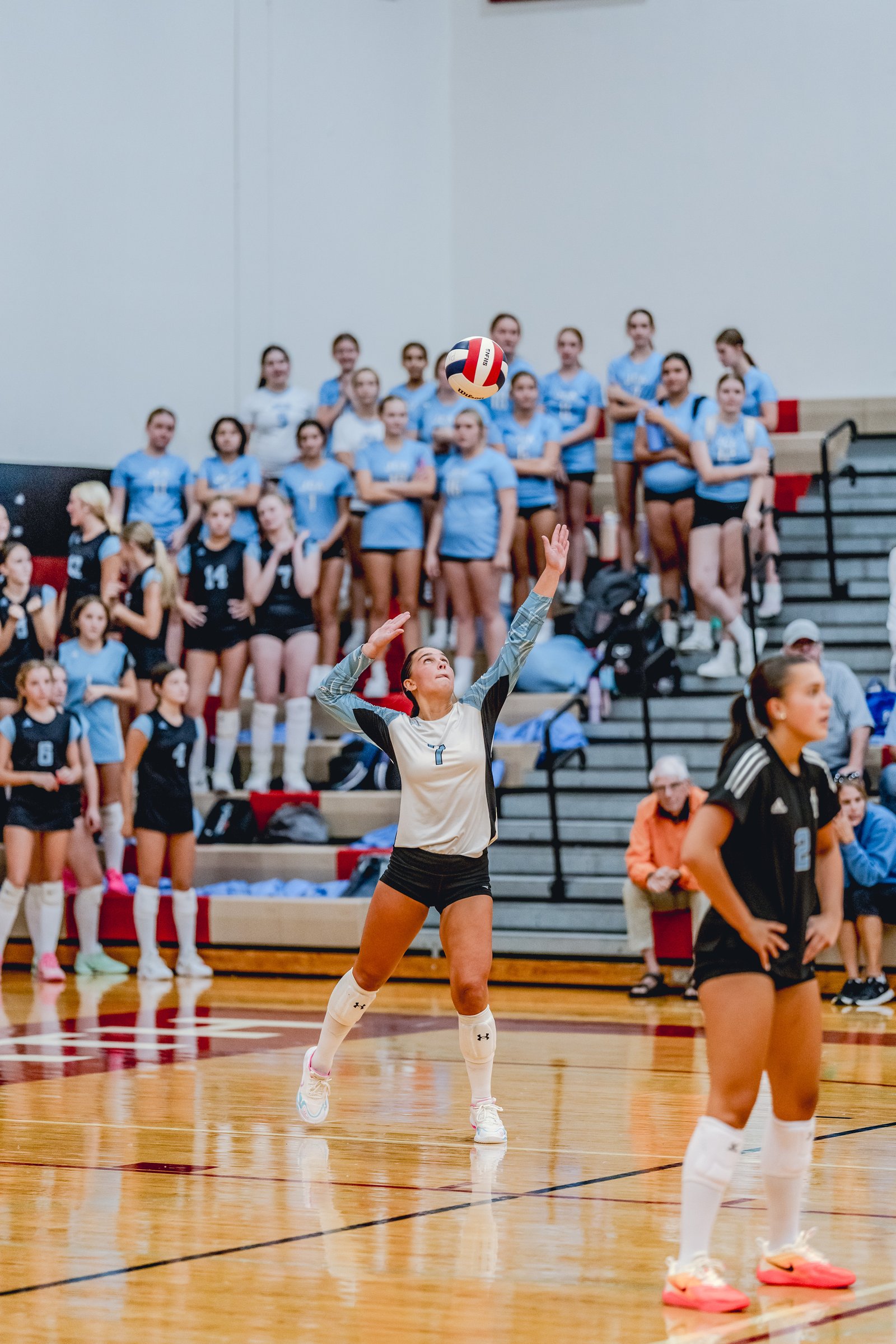 A player in a white and blue top serves a volleyball while teammates in blue watch from the bleachers.