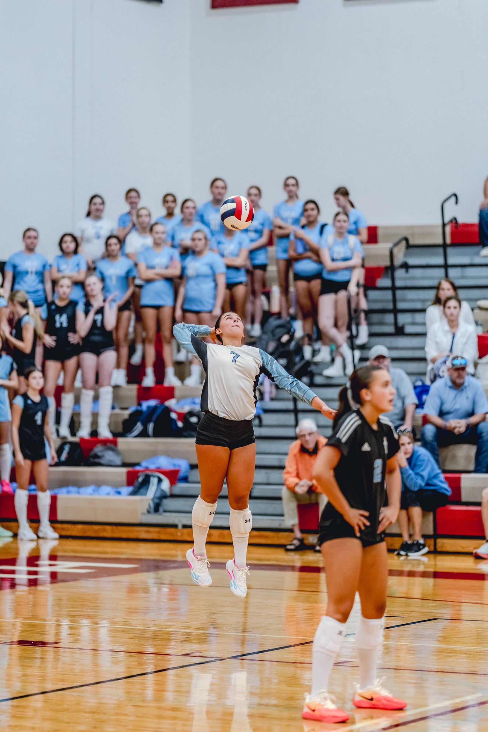 A player in a white and blue jersey with number 7 jumps to serve a volleyball, with a player in a black jersey watching.