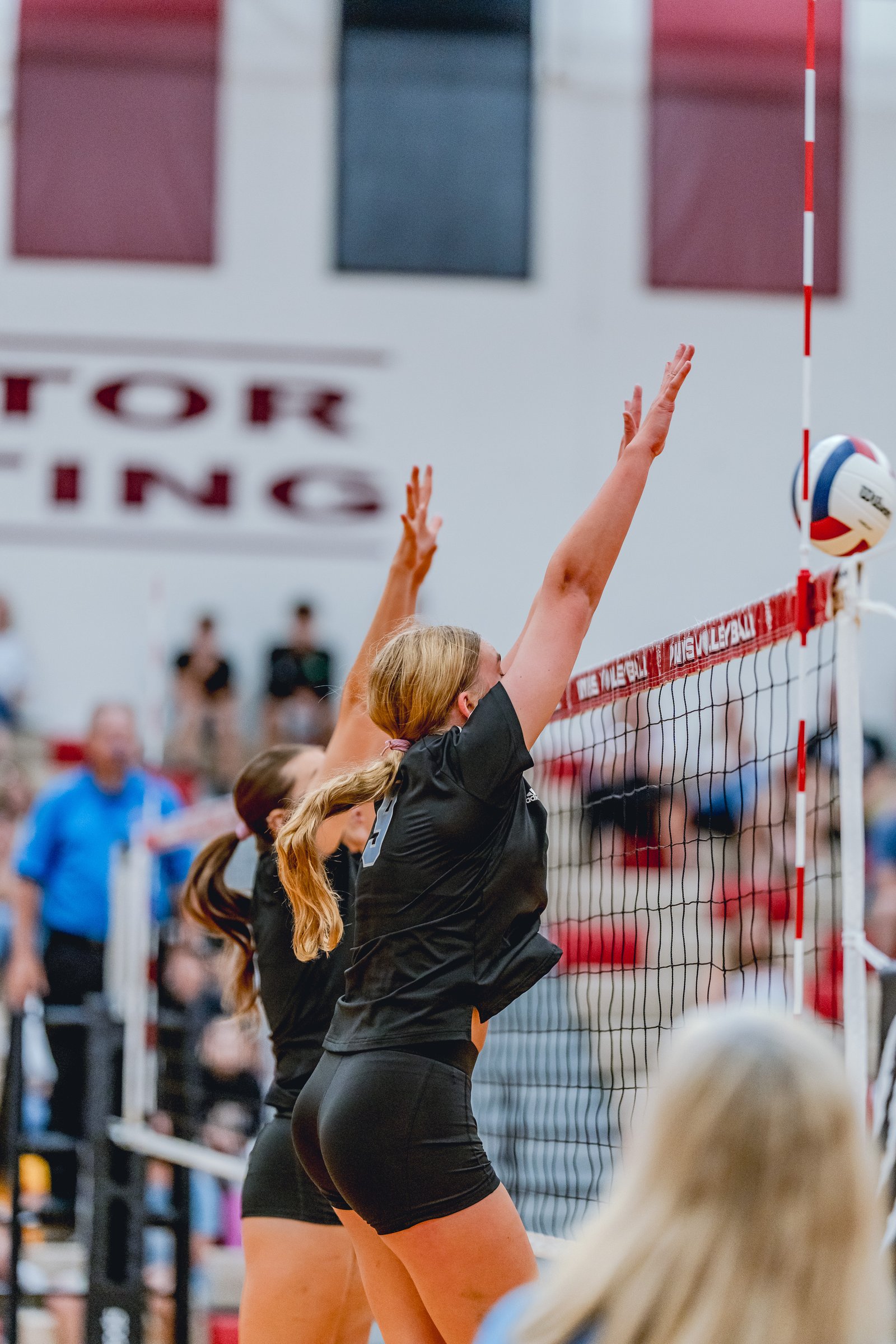Players in black jump at the net during a volleyball game, with a ball in the air.