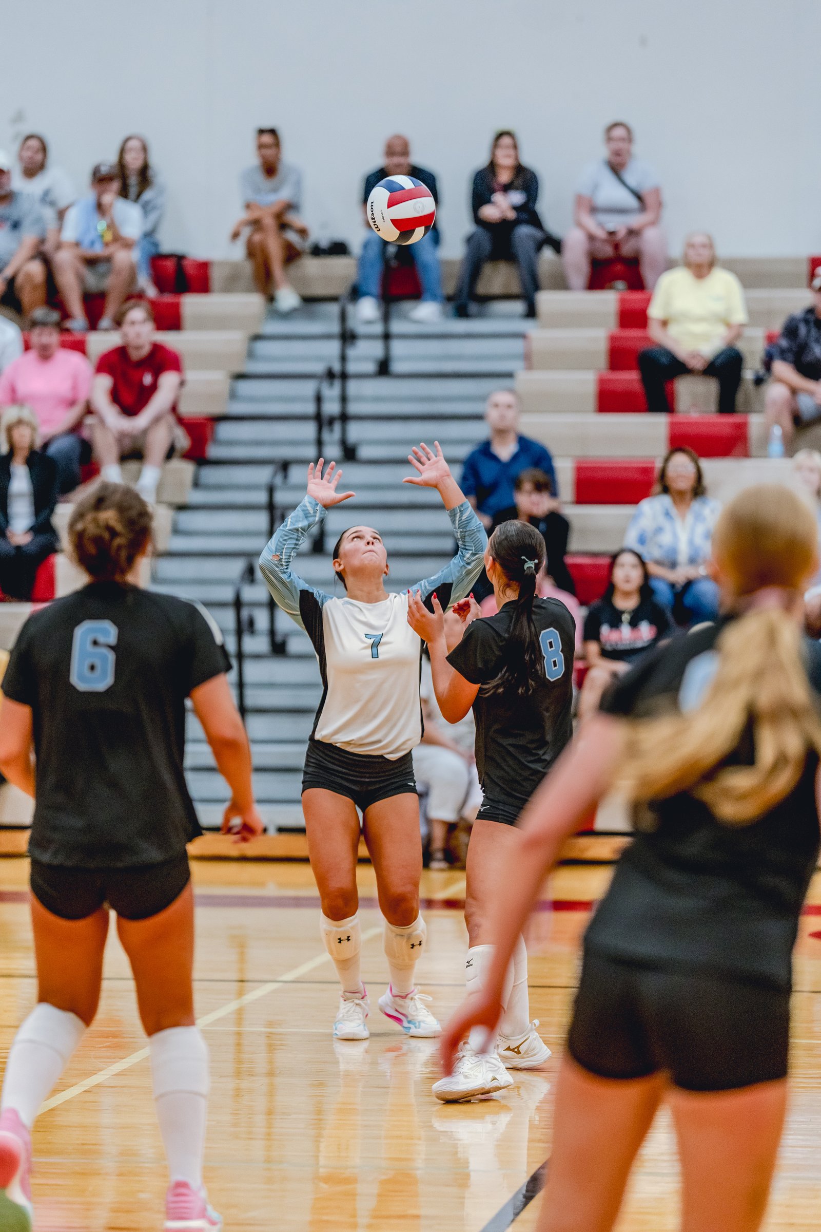 A player in a white and blue jersey with number 7 sets the ball as players in black jerseys with numbers 6 and 8 watch during a volleyball game.