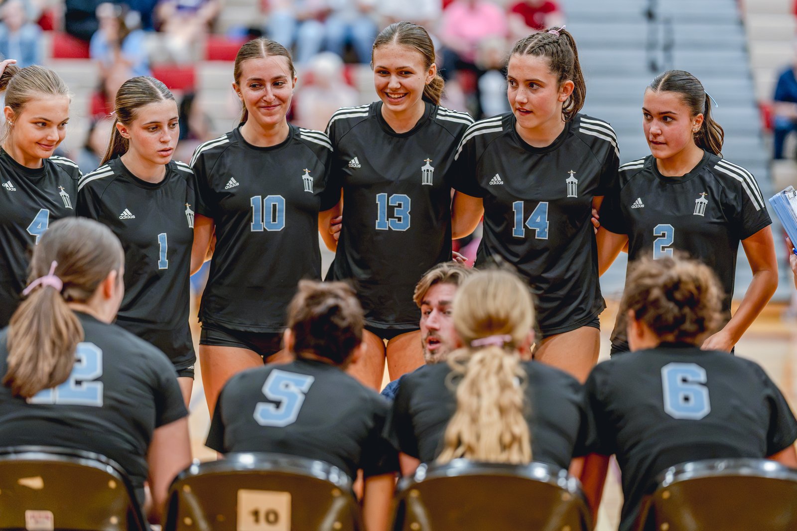 Players in black jerseys with light blue numbers huddle together on the court, with a coach speaking to them.