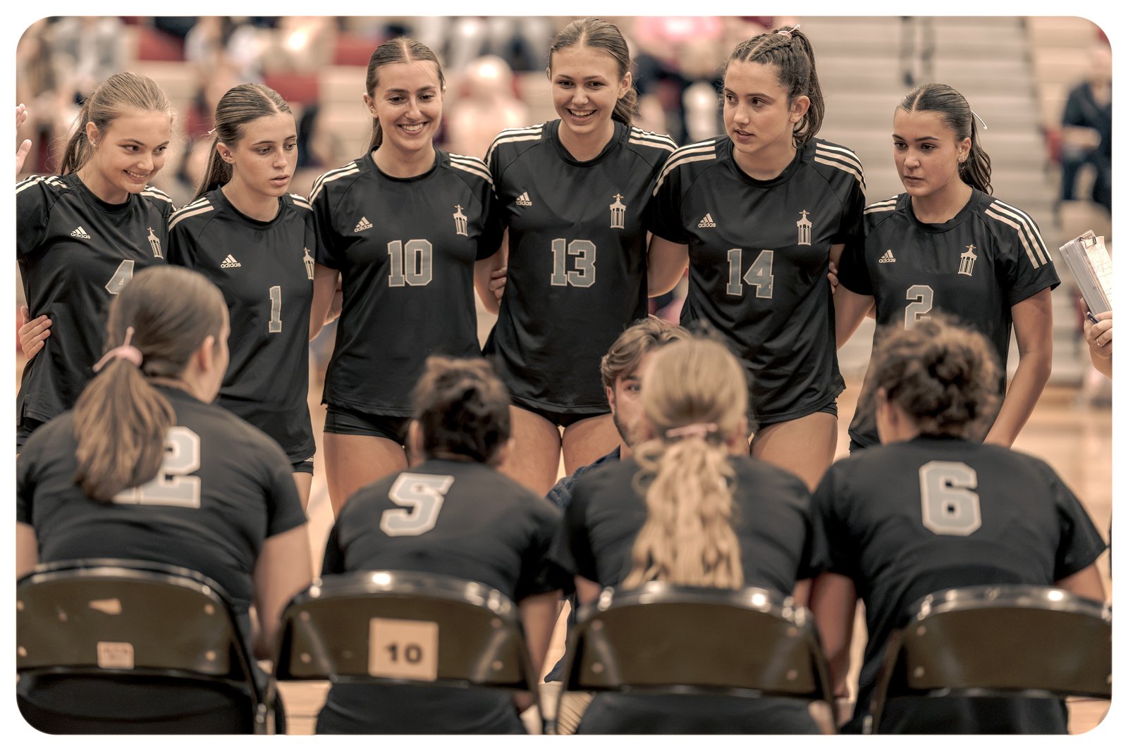 Players in black jerseys with white trim huddle together on the court, some smiling.
