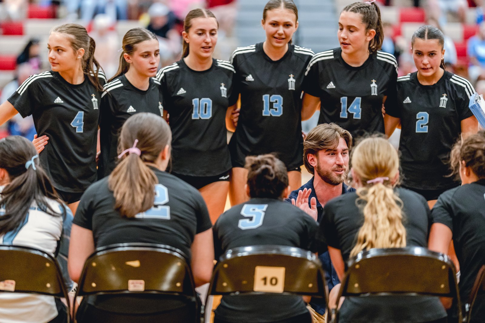 Players in black jerseys with light blue numbers listen to a coach in a blue shirt during a volleyball game.
