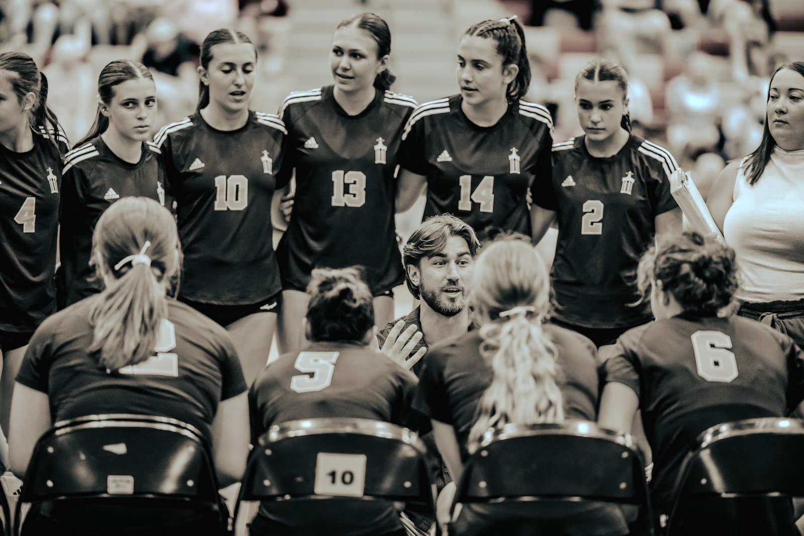 A coach talks to players in dark jerseys with white numbers, including 4, 10, 13, 14, 2, 5, and 6, during a volleyball game.